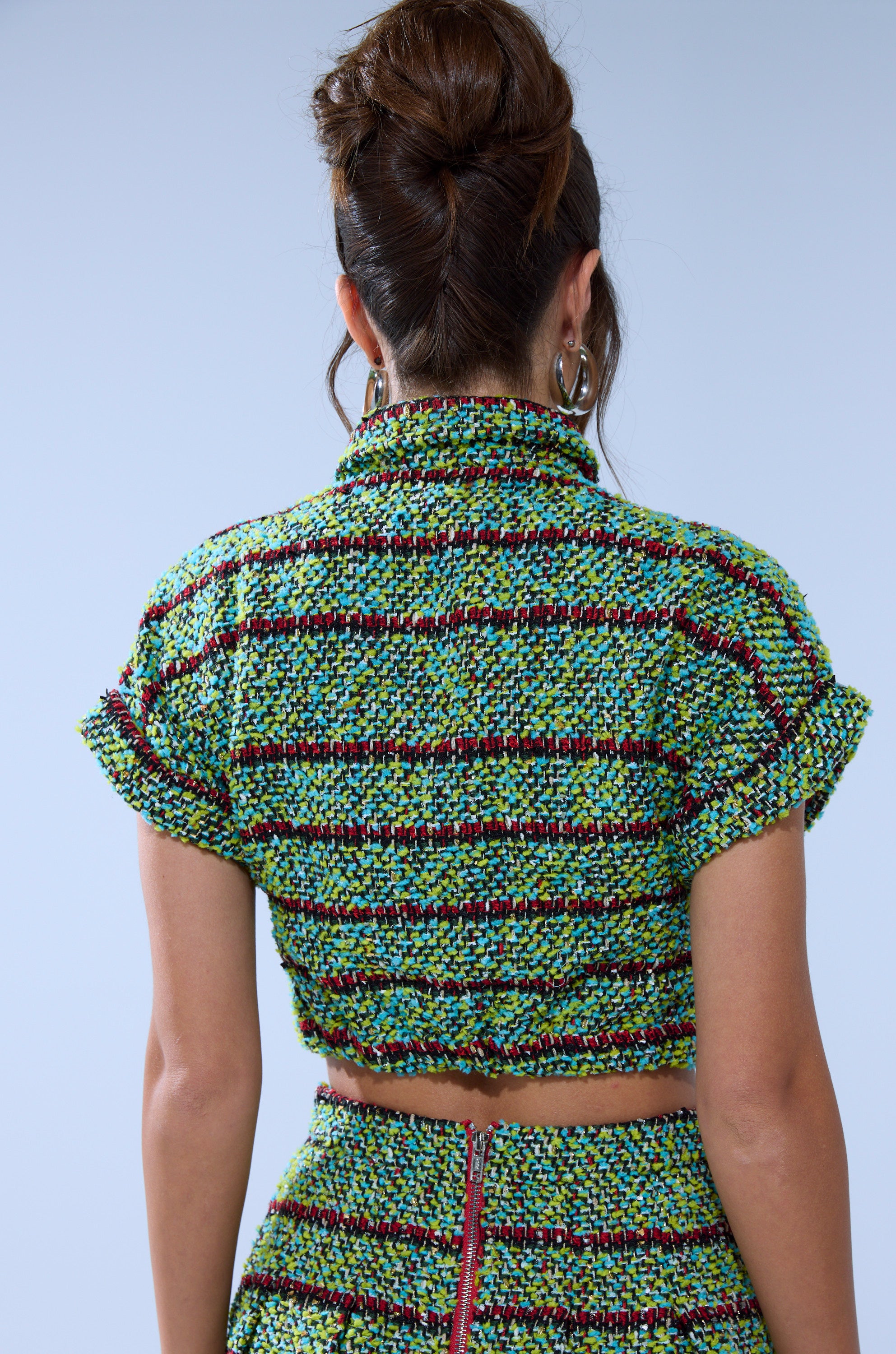Woman with brown hair in an updo wears the IT'S GIVING TWEED BUTTON DOWN—a green, yellow, and red plaid short-sleeved top with matching skirt—shown from the back against a plain light blue background.