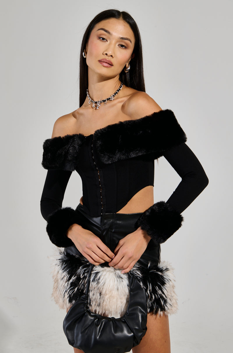 Dressed in the WINTER BAE OFF THE SHOULDER CORSET, a woman stylishly combines her black off-shoulder top featuring faux fur trim with a matching mini skirt adorned with fur accents, creating an ideal holiday party ensemble. She elegantly holds a black and white fur handbag while accessorizing her look with a necklace and earrings. Her long dark hair complements the outfit as she stands against a plain background.