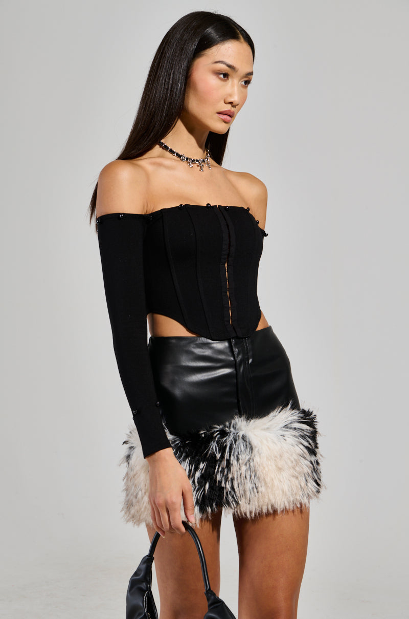 The woman, wearing the WINTER BAE OFF THE SHOULDER CORSET with a chic black leather mini skirt adorned with black and white faux fur trim, carries an elegant handbag. Her long straight hair and necklace add the finishing touches to her look as she gazes to the side against a plain background—ideal for a holiday party.