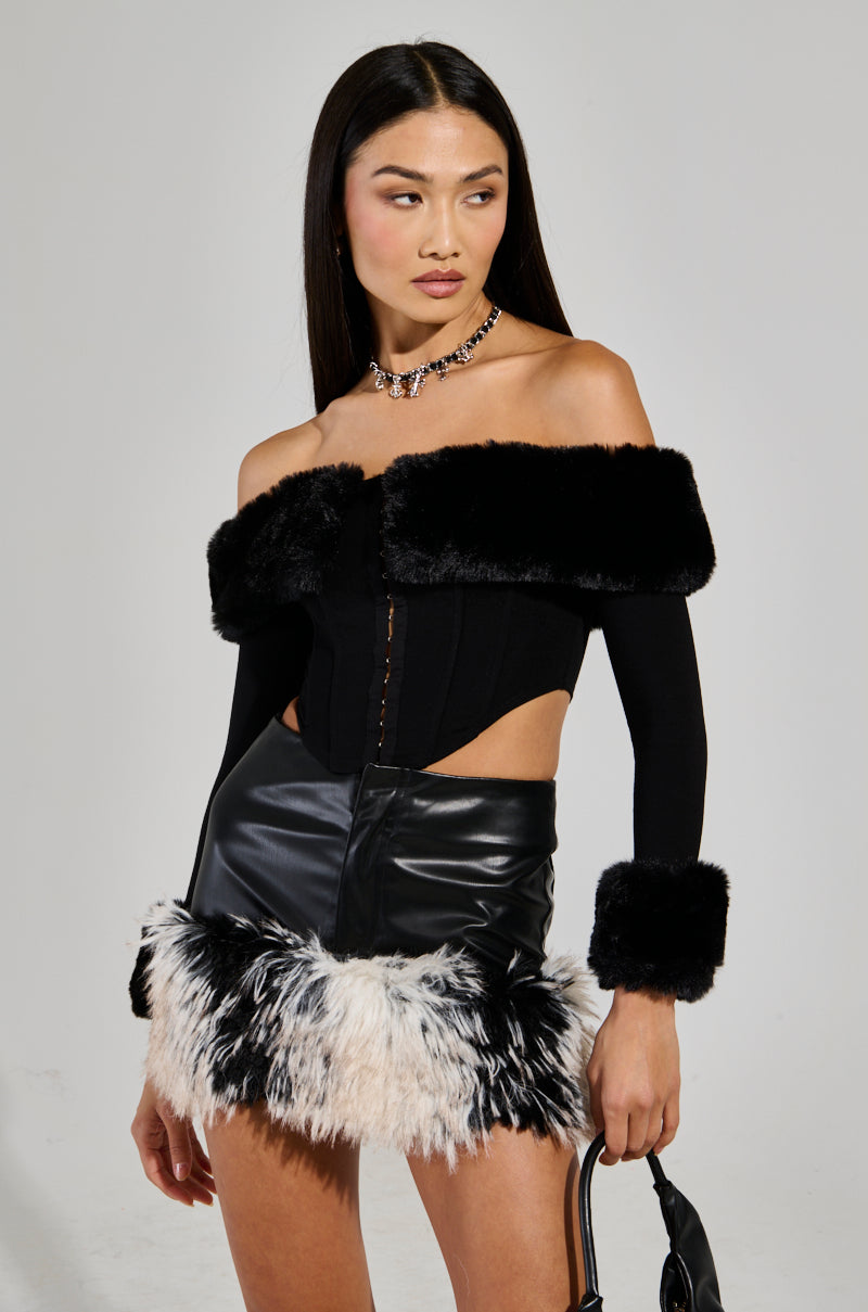 A woman with long dark hair epitomizes a holiday party outfit, wearing the stylish WINTER BAE OFF THE SHOULDER CORSET featuring a luxurious faux fur collar. She pairs it elegantly with a black leather skirt adorned with a furry hem and complements her look with a necklace while holding a small handbag against the plain backdrop.