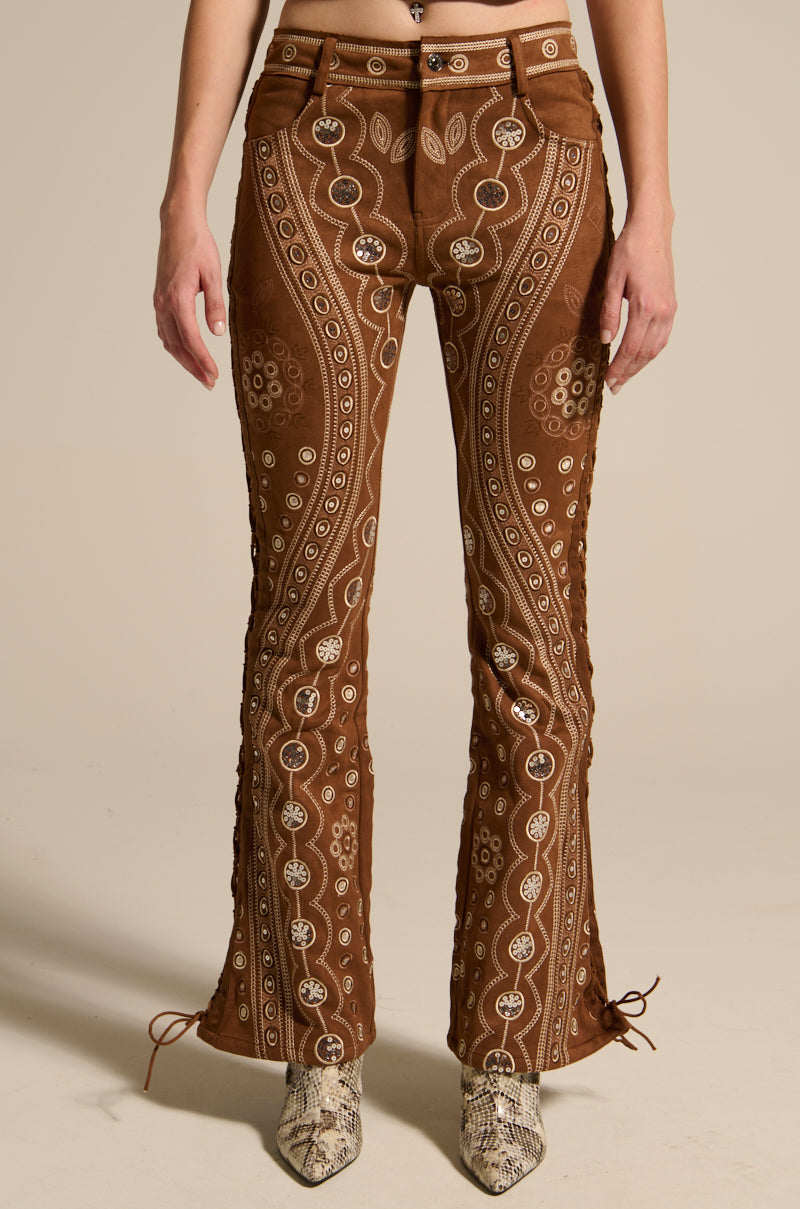 Someone is wearing the TEXAS FOREVER PANT, Western-inspired brown flared pants with intricate white paisley and circular patterns. The faux suede pants have lace-up detailing on the sides and pair perfectly with brown and beige snake-patterned shoes against a plain beige background.