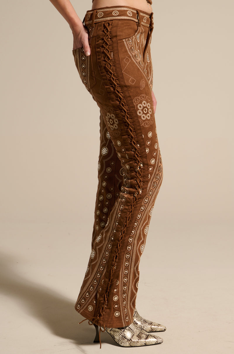 A person wears the TEXAS FOREVER PANT, Western-inspired faux suede pants with white patterns and lace-up sides, paired with pointed snakeskin boots, standing against a plain light background.