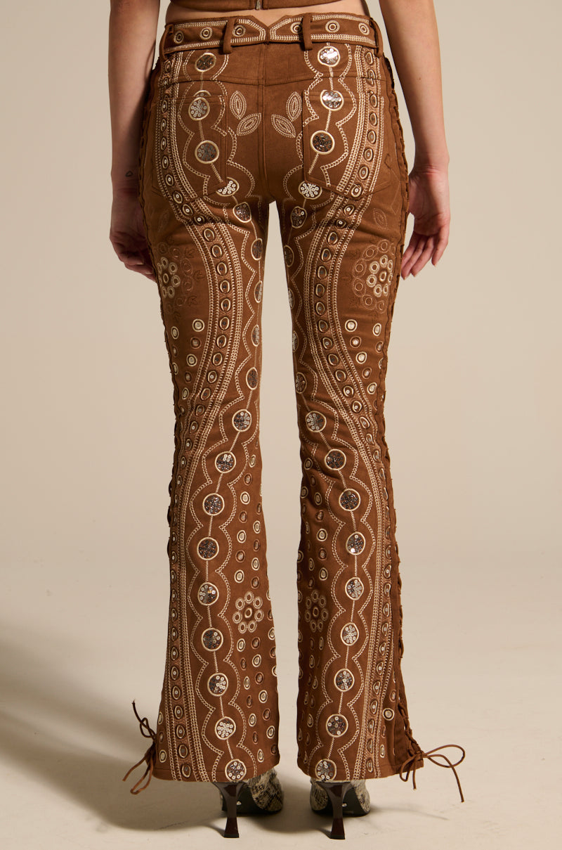 Someone wears TEXAS FOREVER PANT, brown bell-bottoms with white and beige embroidery of circles and floral motifs. Made from faux suede, they have side lace-up details and are paired with heeled boots against a plain background.