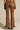 Someone wears TEXAS FOREVER PANT, brown bell-bottoms with white and beige embroidery of circles and floral motifs. Made from faux suede, they have side lace-up details and are paired with heeled boots against a plain background.
