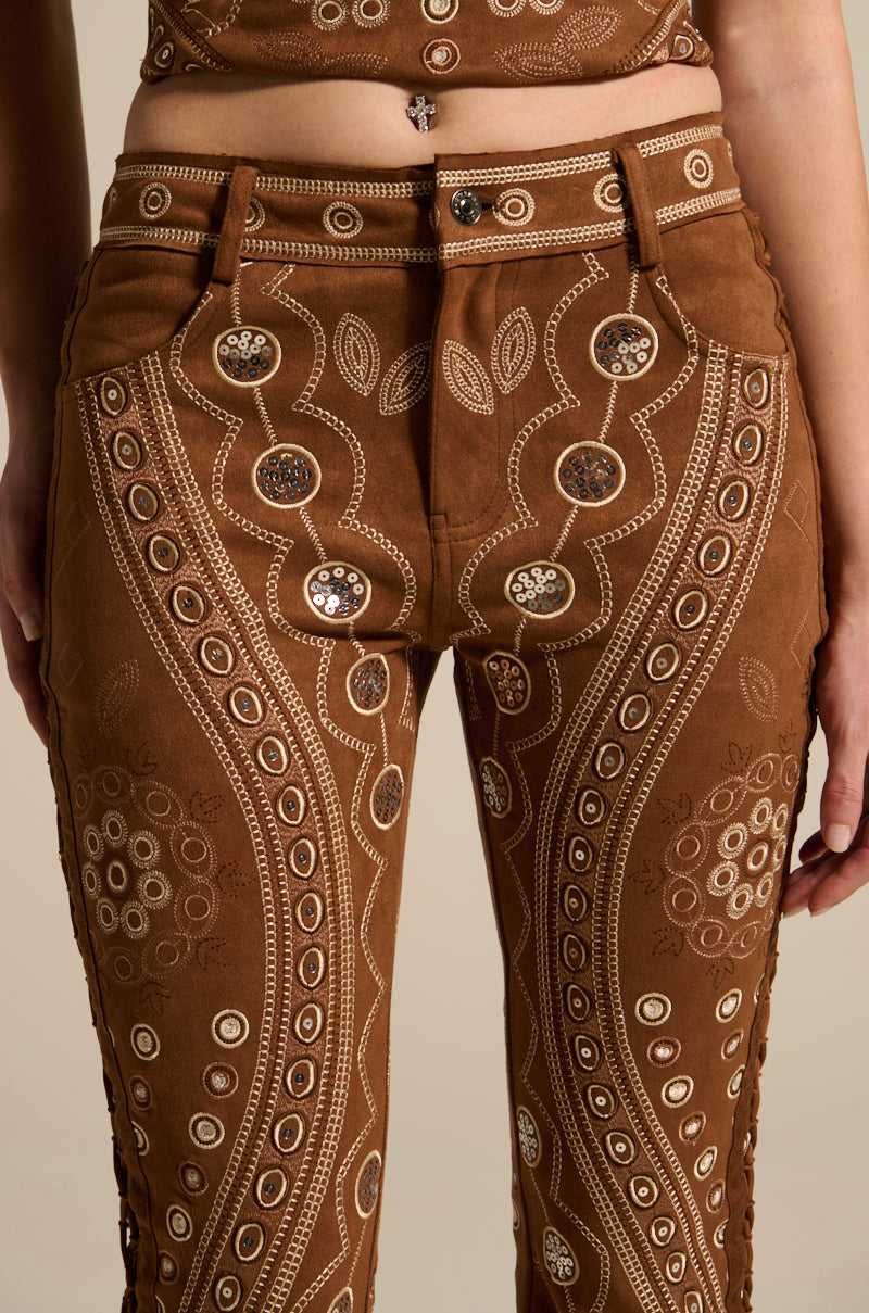 The TEXAS FOREVER PANT, crafted from faux suede with intricate beige embroidery and circular metal embellishments, features a swirling design and subtle lace-up detailing. The image highlights the craftsmanship on the lower abdomen and legs of these brown, Western-inspired pants.