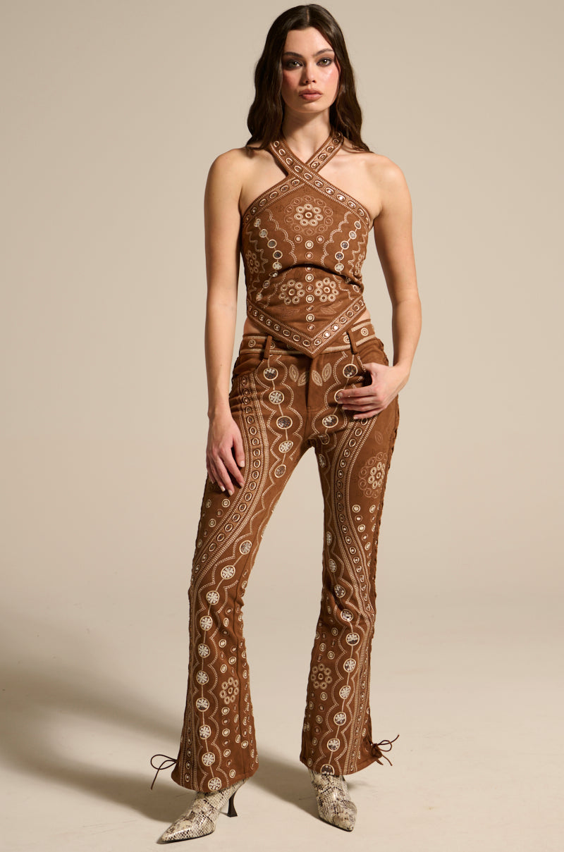 A woman is showcasing the TEXAS FOREVER HALTER TOP, a brown, patterned jumpsuit with a Western halter neck and detailed cowgirl embroidery in white. She's styled with heeled boots against a plain beige backdrop.