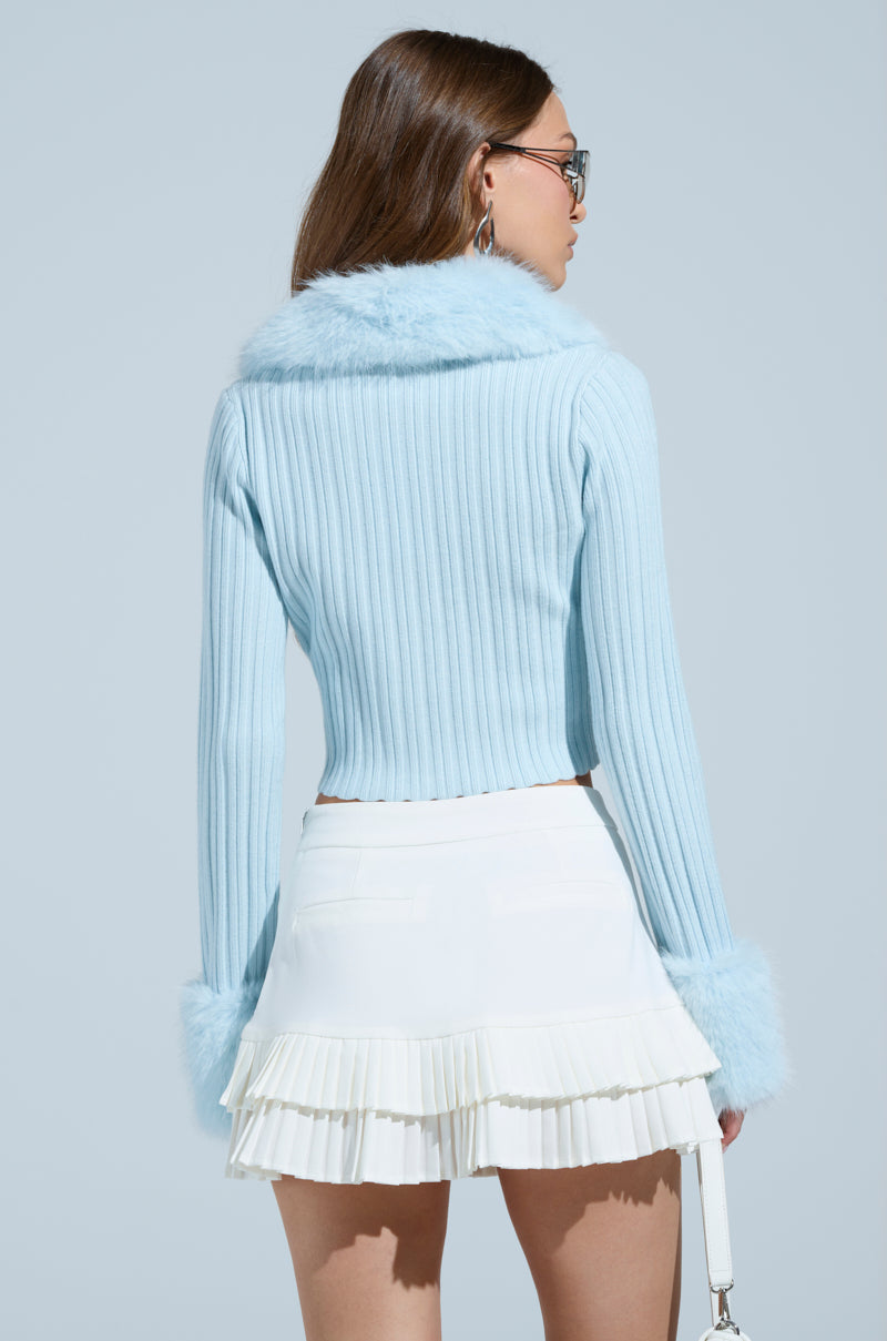 A person with long brown hair and glasses is shown from the back, wearing an Angel Eyes Fur Trim Cardigan in blue, featuring a faux fur collar and fluffy cuffs, elegantly paired with a white pleated skirt against a serene light blue background.