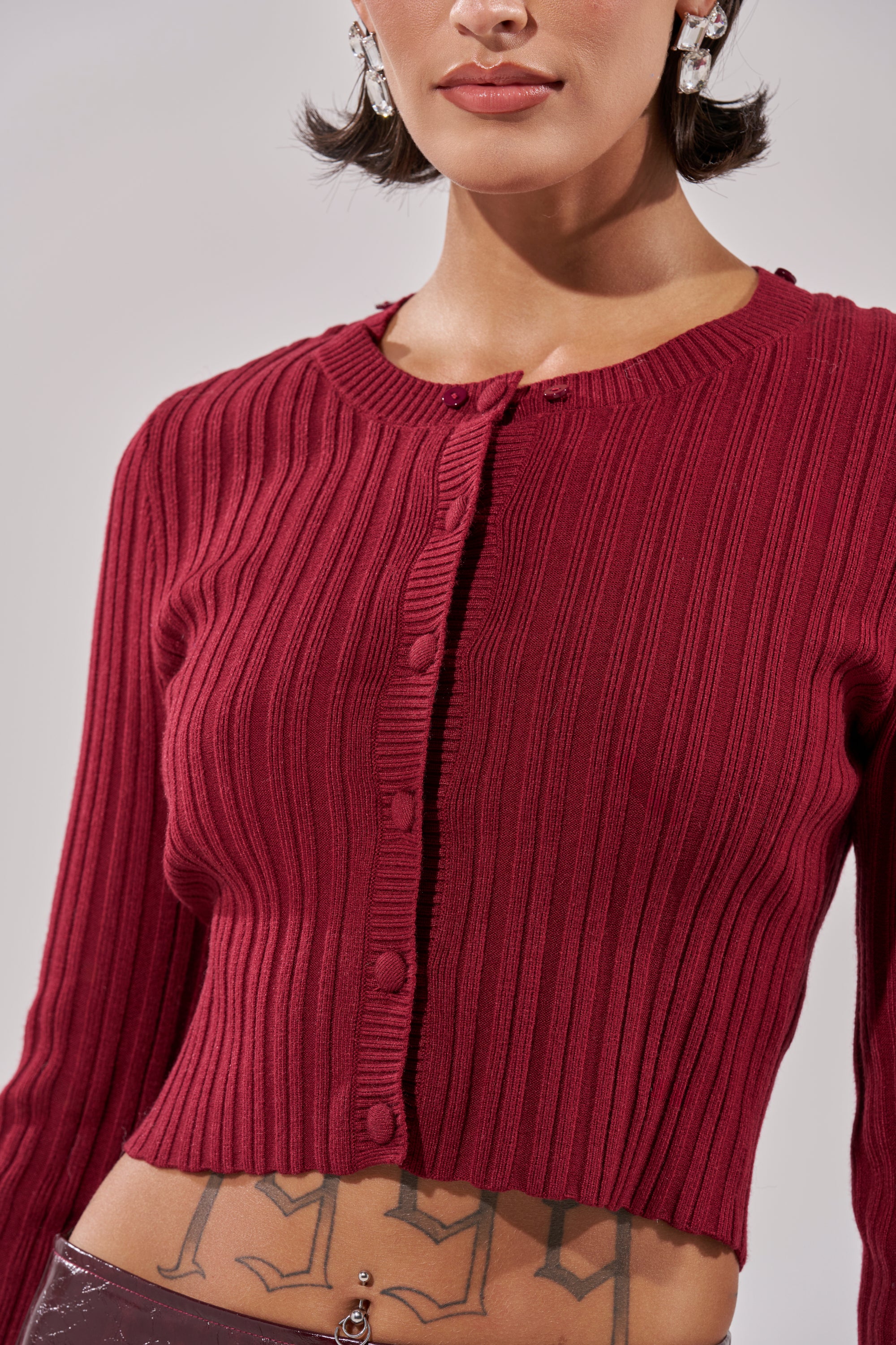 A woman with short dark hair, visible earrings, and a bold numerical tattoo on her lower abdomen wears the EASE MY MIND CARDIGAN WITH FUR TRIM—a ribbed knit, long-sleeve burgundy cardigan with buttons. 11