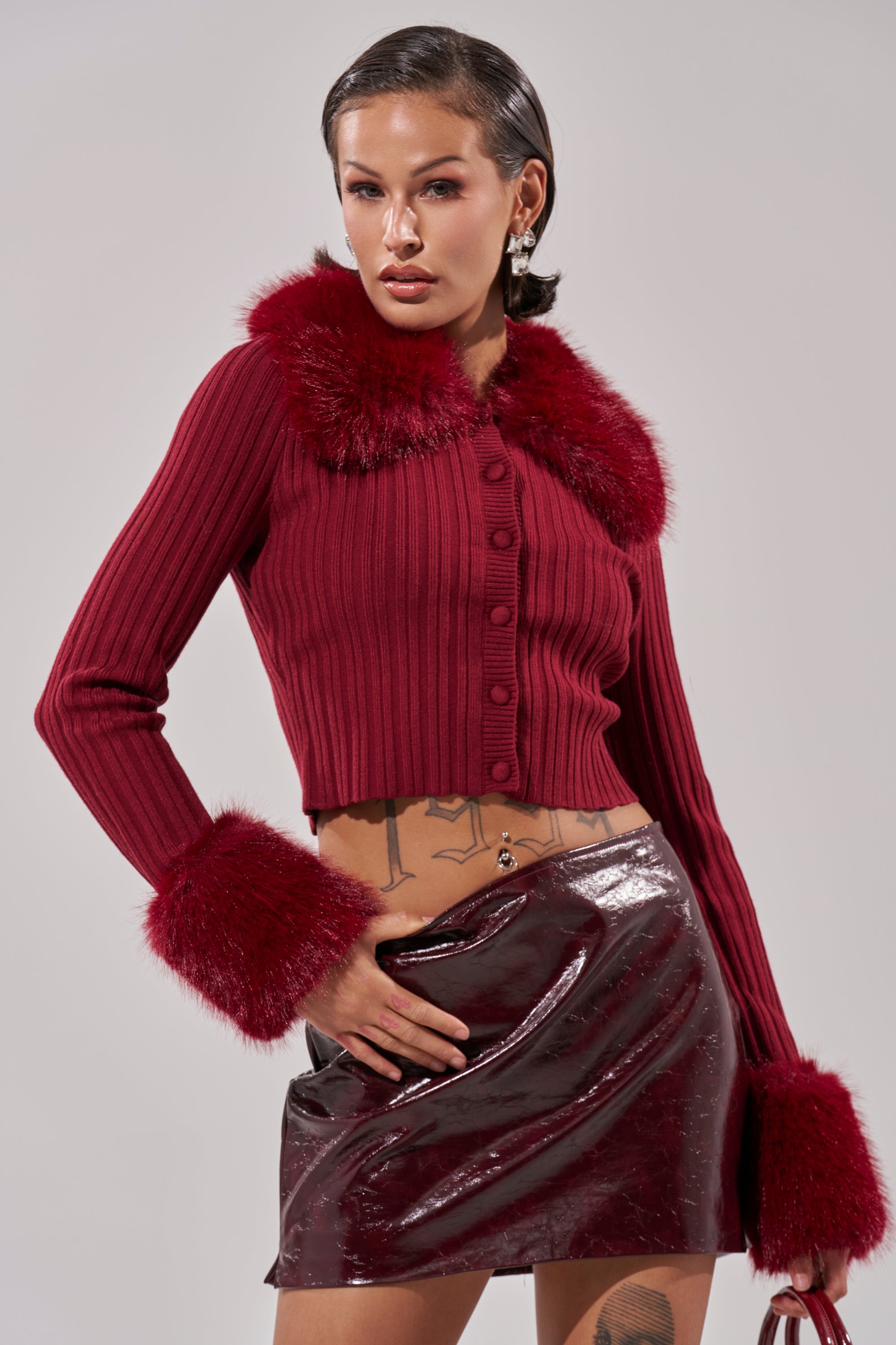 A woman poses confidently in the EASE MY MIND CARDIGAN WITH FUR TRIM, a red ribbed knit with faux fur collar and cuffs, styled with a shiny burgundy mini skirt. She has short, slicked-back hair and visible tattoos against a neutral background. 2