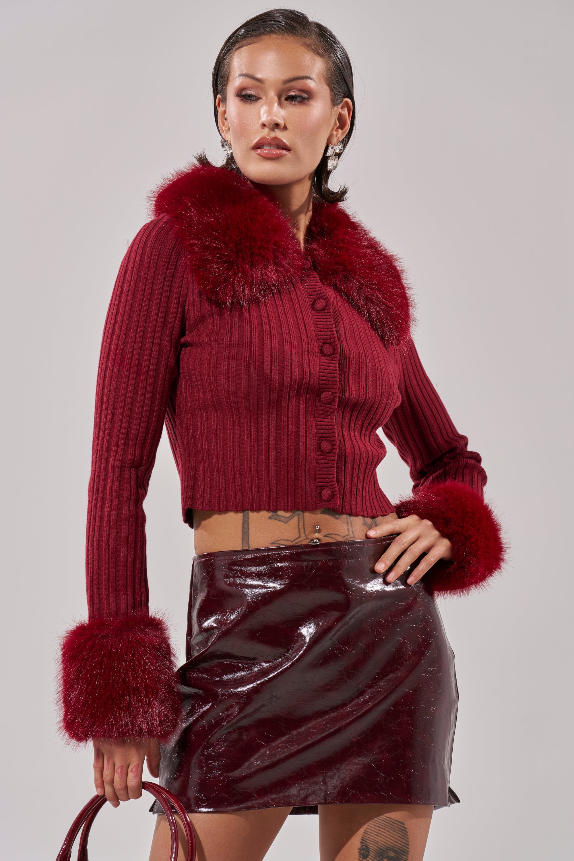 A woman poses confidently against a plain light background wearing the EASE MY MIND CARDIGAN WITH FUR TRIM and a shiny burgundy mini skirt. The ribbed knit cardigan features faux fur at the collar and cuffs for a chic look. 3