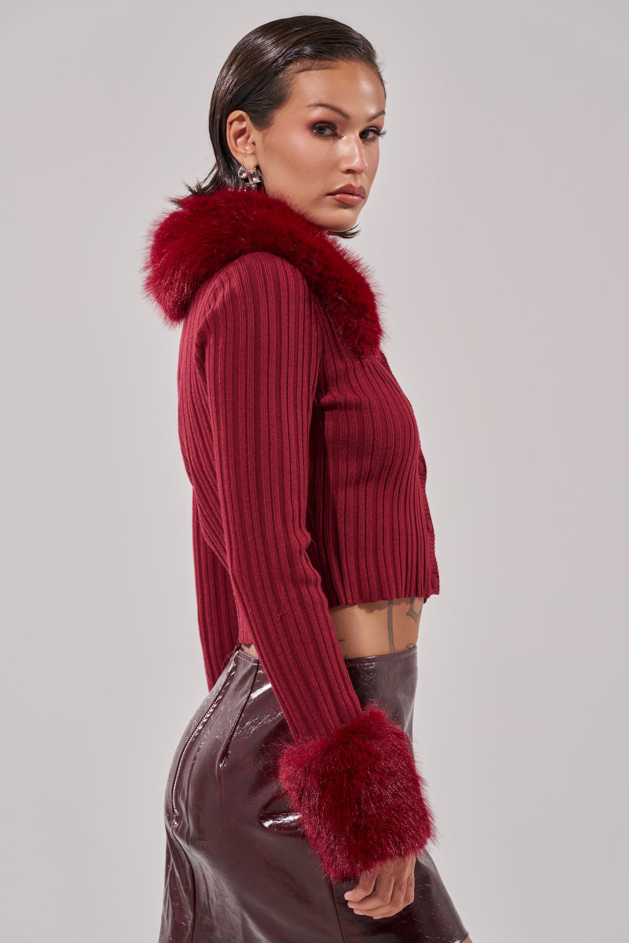 A woman with short dark hair models the EASE MY MIND CARDIGAN WITH FUR TRIM, a ribbed knit cropped style featuring faux fur collar and cuffs, paired with a shiny dark burgundy skirt against a light gray background. 4