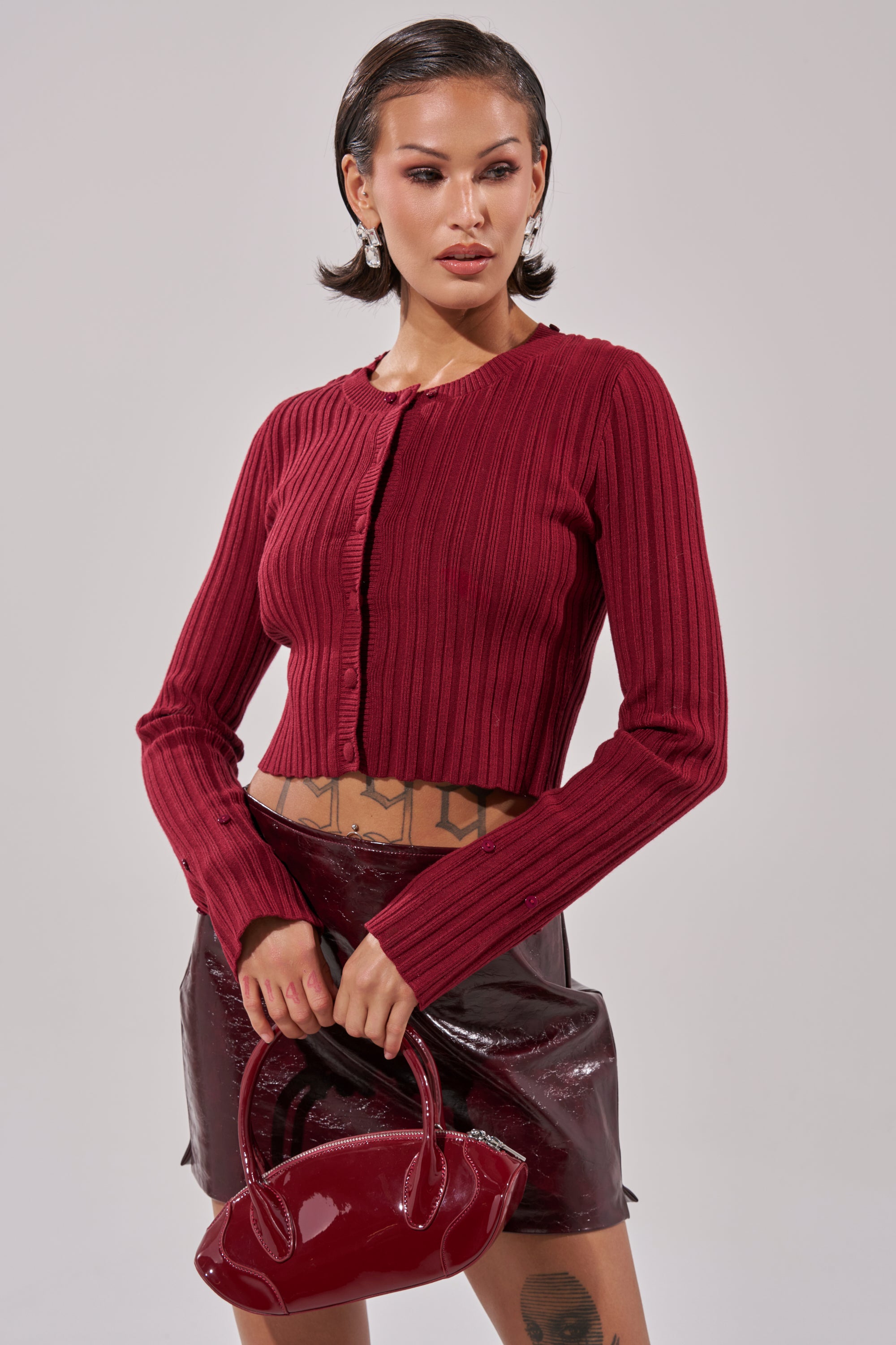 A woman wears the EASE MY MIND CARDIGAN WITH FUR TRIM—a ribbed, long-sleeve red cropped cardigan—paired with a shiny dark red mini skirt and matching glossy handbag. She has short, slicked-back hair and visible tattoos above her skirt. 5