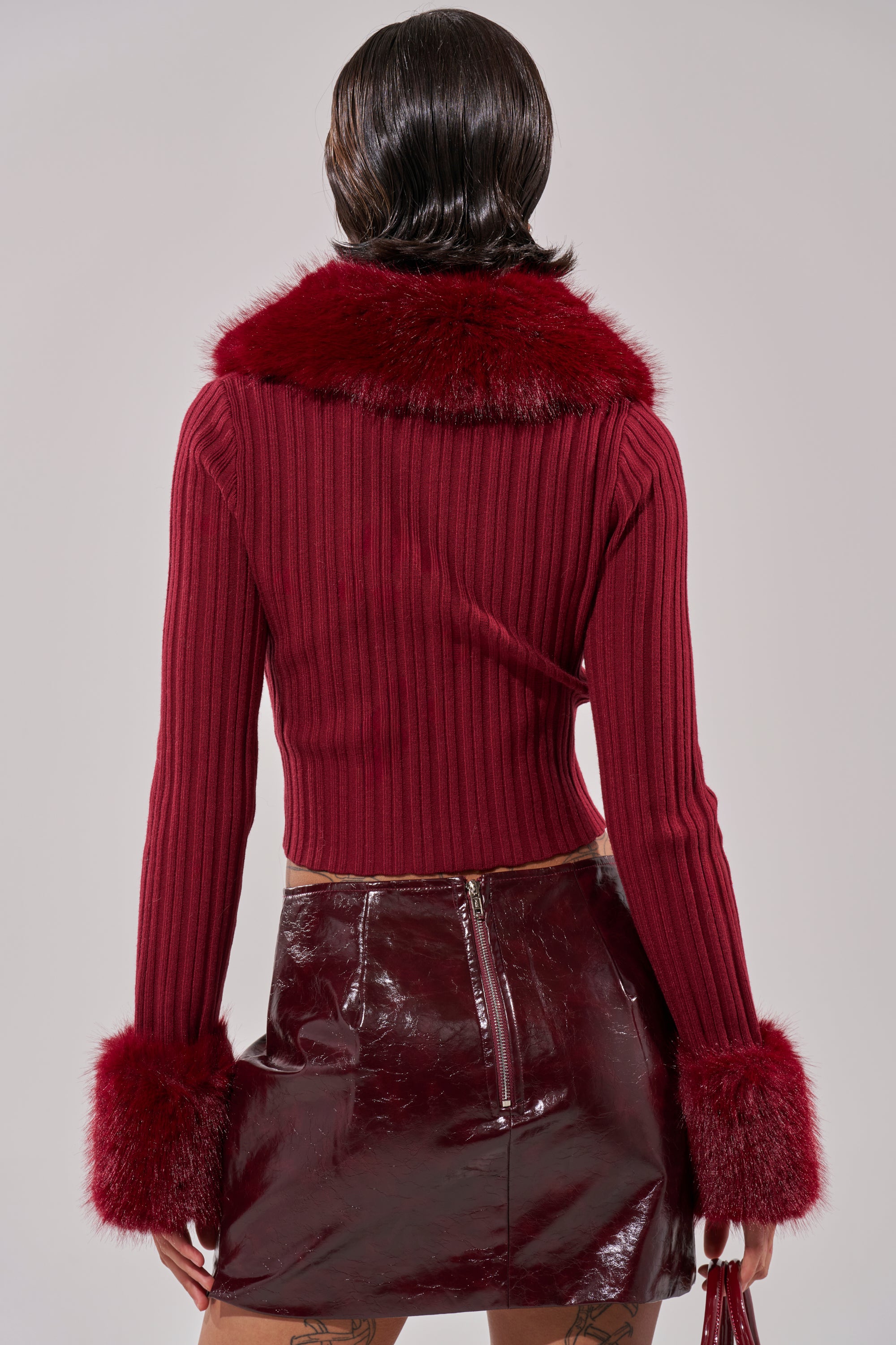 A person with short, dark hair is seen from the back, wearing the EASE MY MIND CARDIGAN WITH FUR TRIM and a shiny, dark red vinyl skirt with a visible back zipper. 6