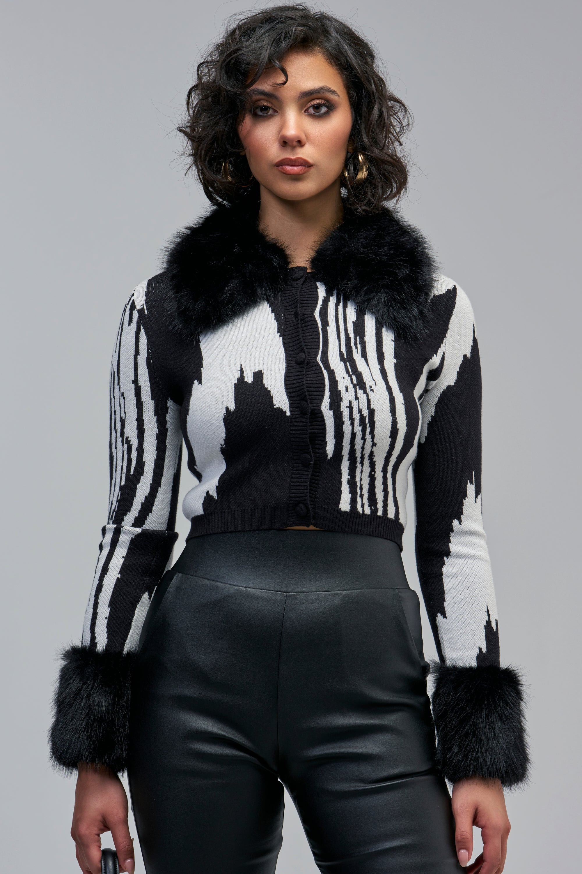 A woman wears the EASE MY MIND CARDIGAN WITH FUR TRIM IN BLACK WHITE, a cropped sweater with an abstract print, black faux fur collar, and cuffs.