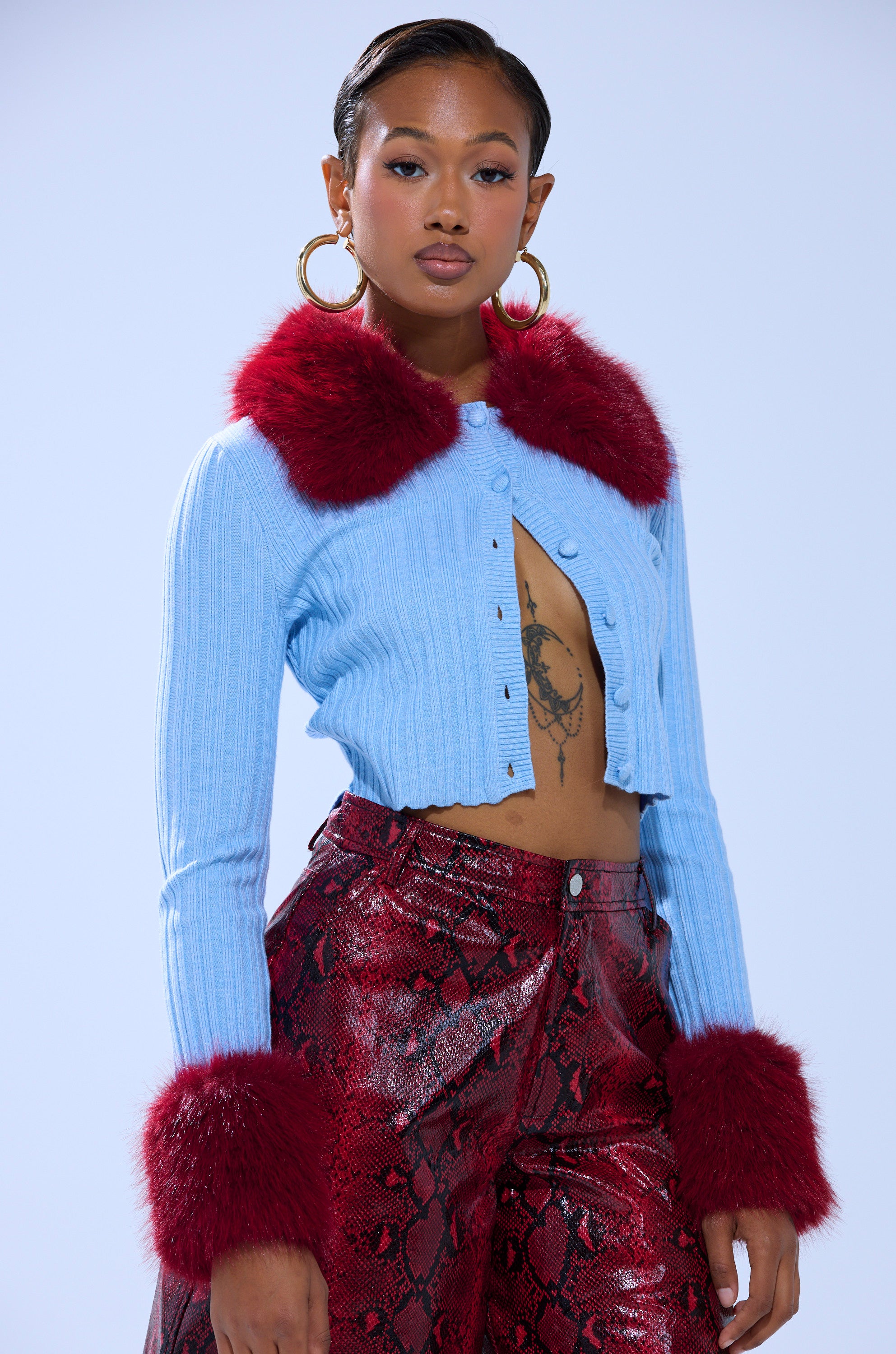 A woman wears the EXTRA IS MY MIDDLE NAME CARDIGAN WITH FUR, a light blue cropped knit featuring faux fur trim on the collar and cuffs, paired with high-waisted red snakeskin pants and large hoop earrings that showcase her confident style.