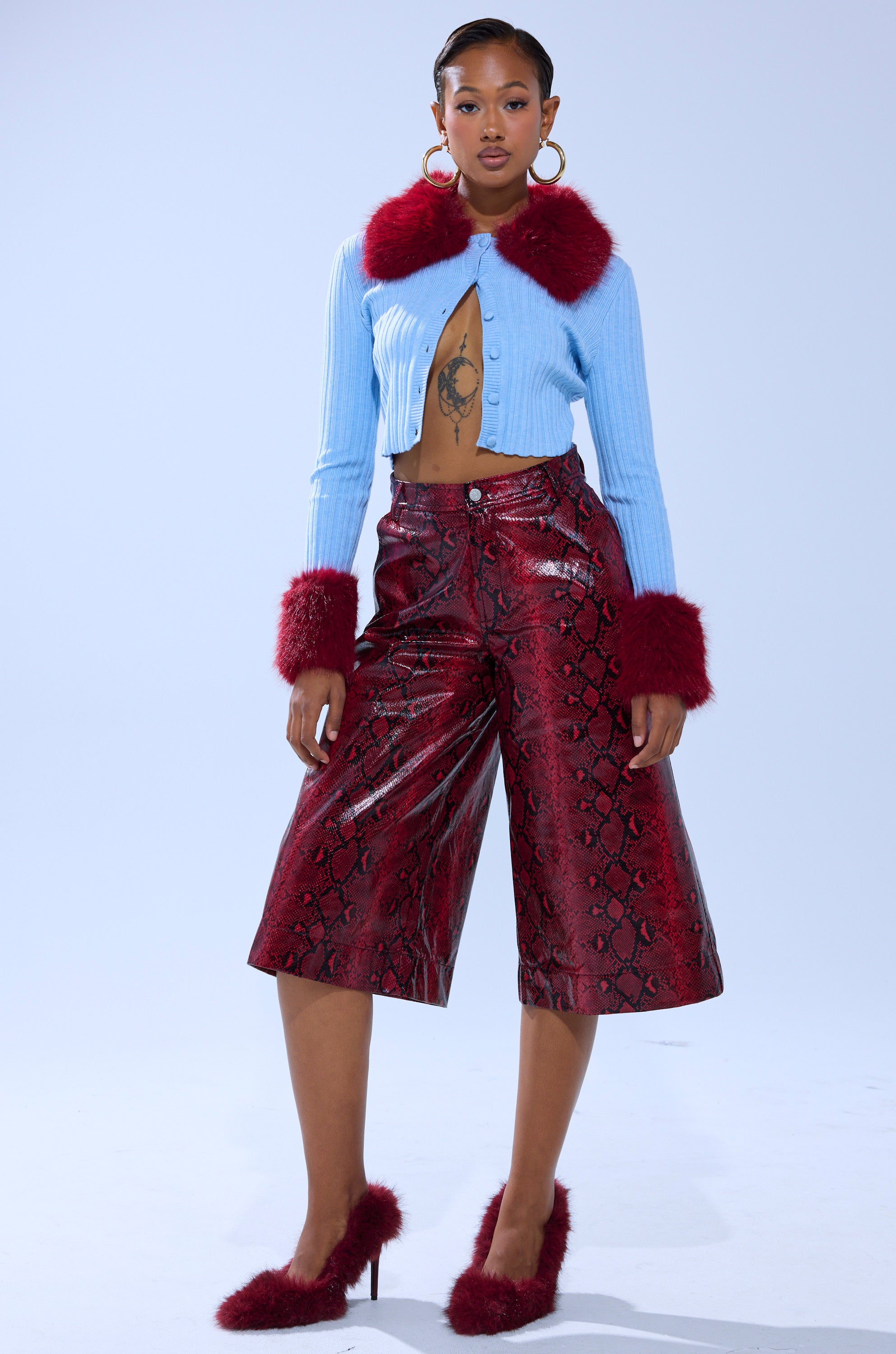A woman poses stylishly in the EXTRA IS MY MIDDLE NAME CARDIGAN WITH FUR, featuring red faux fur trim. She pairs it with red snakeskin culottes, matching furry heels, large hoop earrings, and displays a visible chest tattoo.