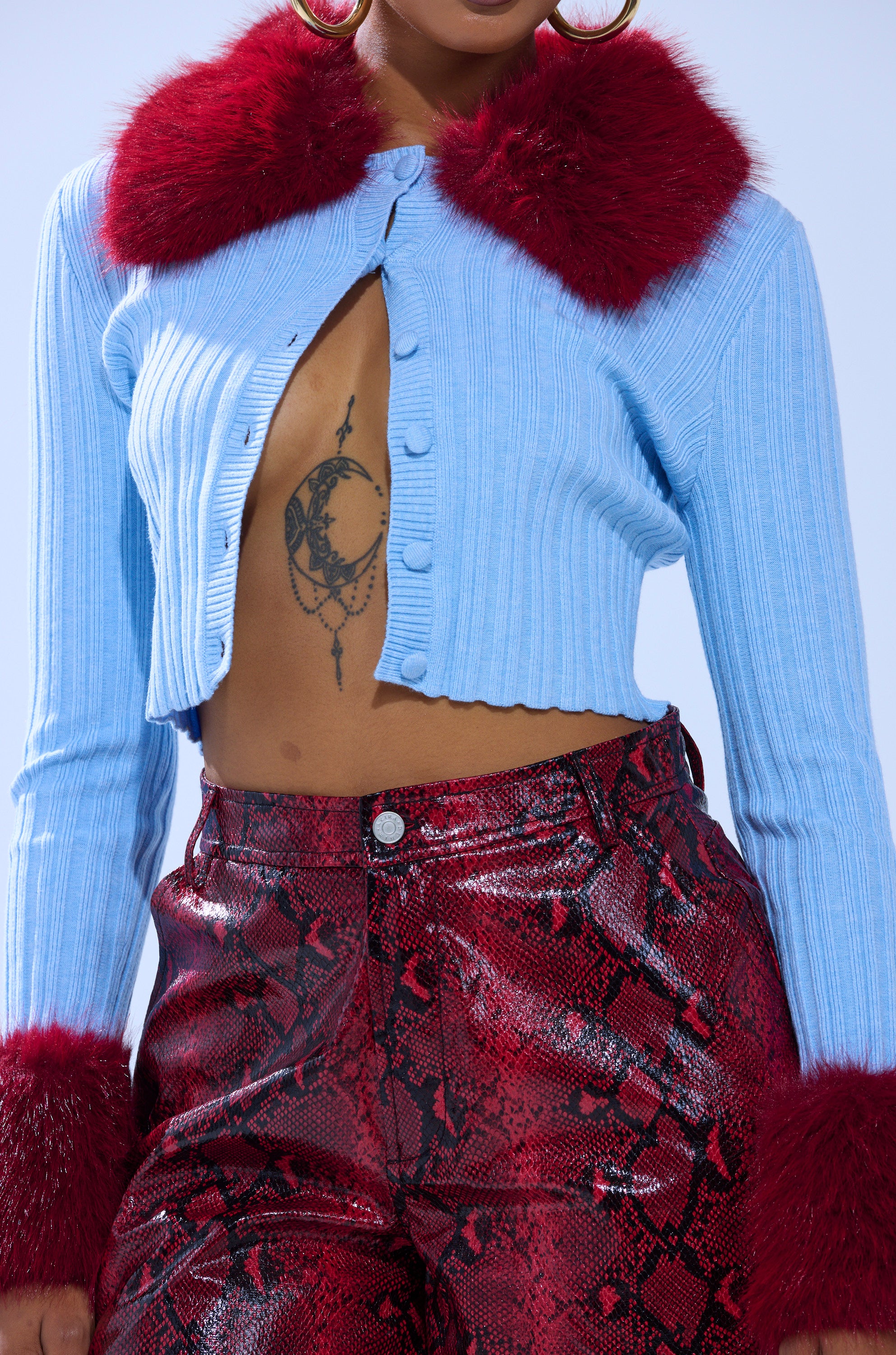 A person wears the EXTRA IS MY MIDDLE NAME CARDIGAN WITH FUR in light blue with red faux fur collar and cuffs, paired with shiny red snakeskin-patterned pants. A crescent moon tattoo is partly visible on their chest.