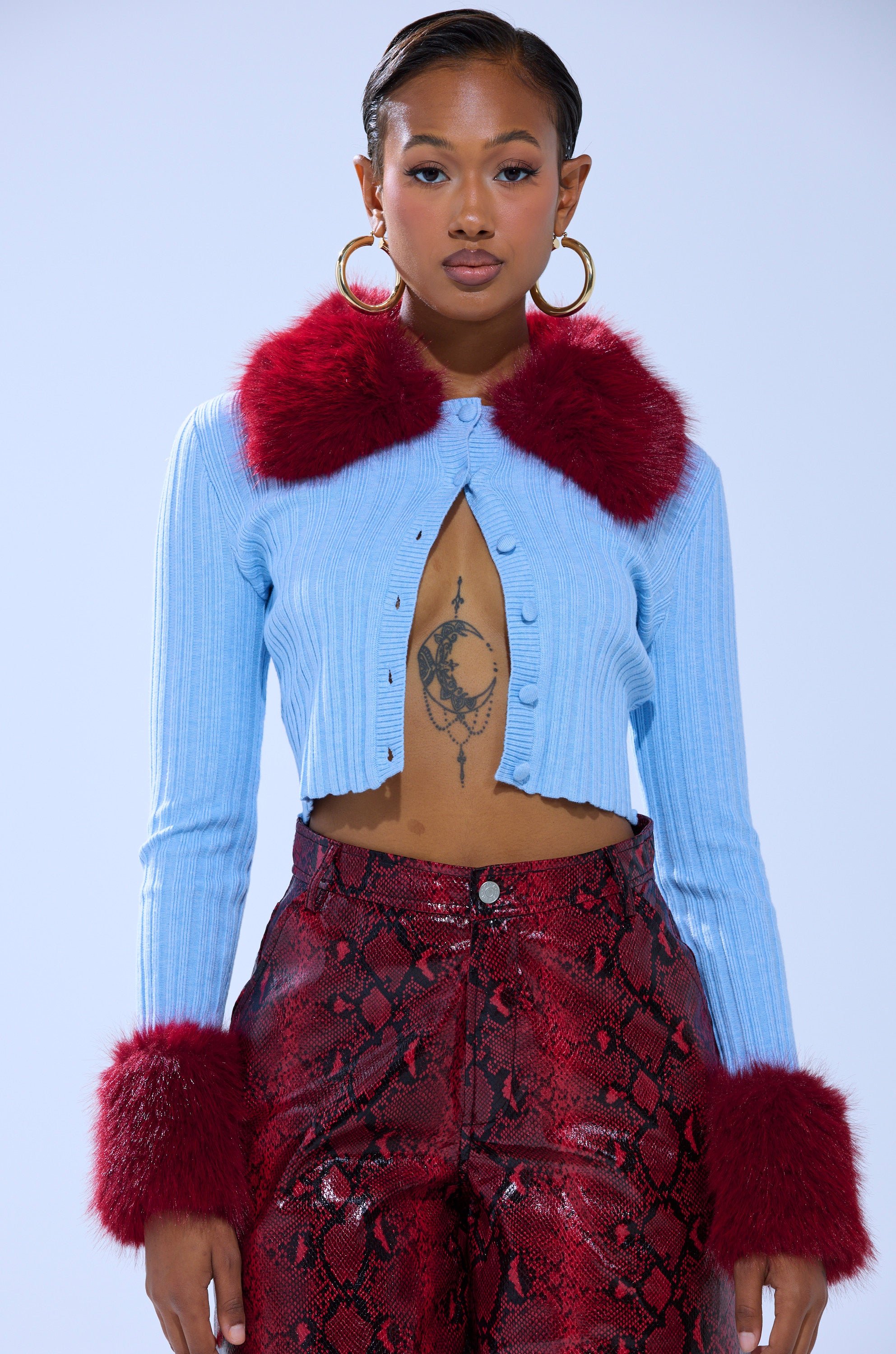 A woman faces forward in the EXTRA IS MY MIDDLE NAME CARDIGAN WITH FUR—light blue with red faux fur collar and cuffs—paired with red snakeskin pants, large hoop earrings, and a crescent moon and sun chest tattoo.