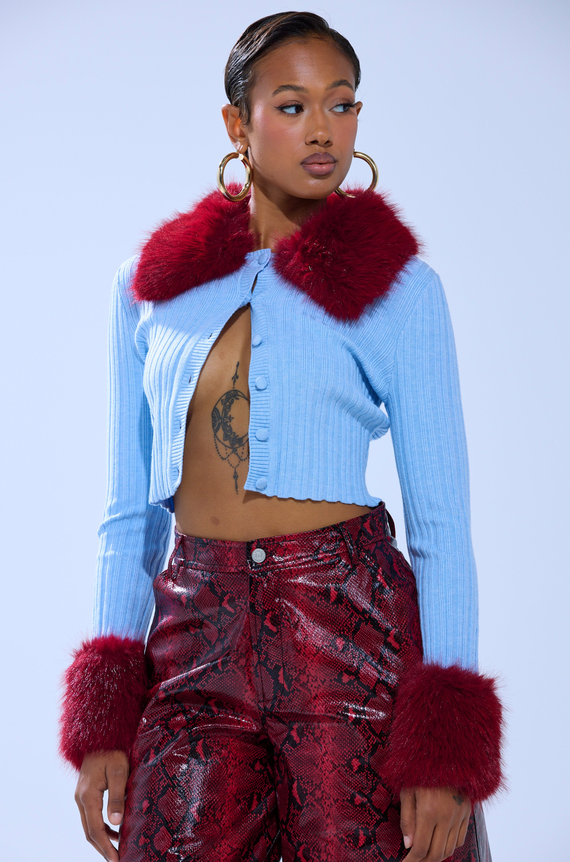 A woman with slicked-back hair wears large hoop earrings and the EXTRA IS MY MIDDLE NAME CARDIGAN WITH FUR, styled with shiny red snake-print pants, posing confidently against a pale background.