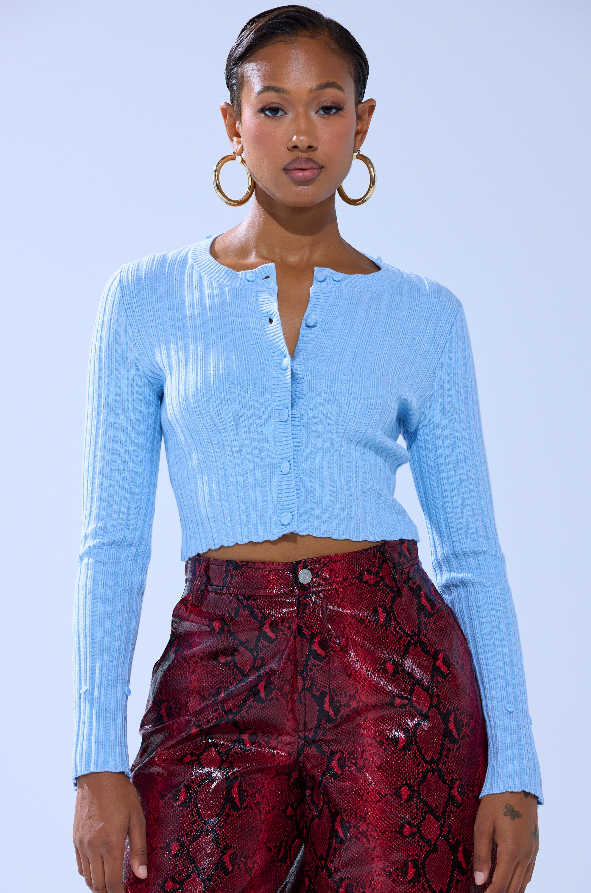 A woman wears the EXTRA IS MY MIDDLE NAME CARDIGAN WITH FUR, large hoop earrings, and high-waisted red snakeskin-print pants, standing against a plain light blue background.