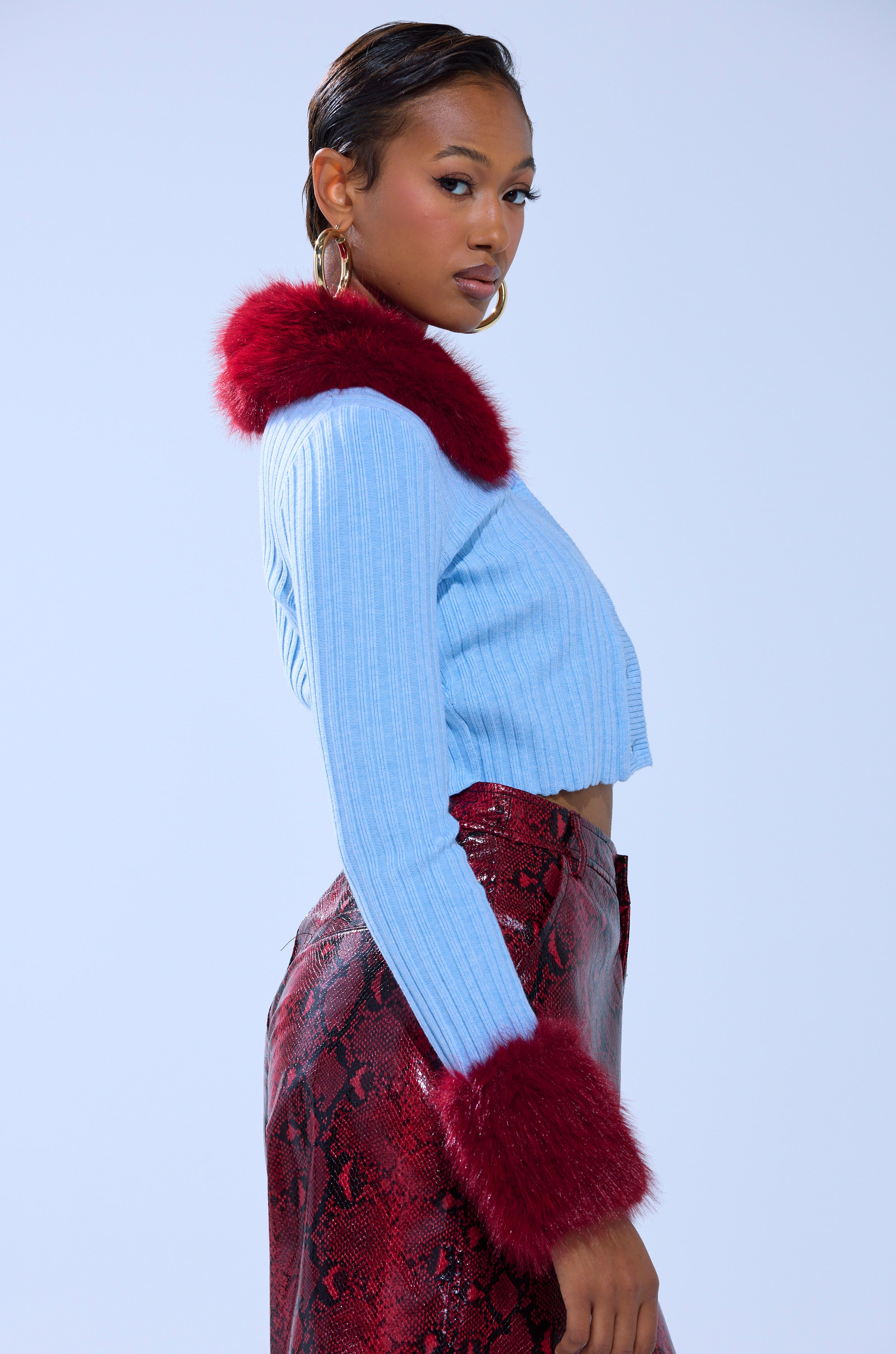 A woman poses sideways in the EXTRA IS MY MIDDLE NAME CARDIGAN WITH FUR, a blue ribbed knit crop with red faux fur trim, paired with high-waisted red snakeskin pants against a plain light blue background.
