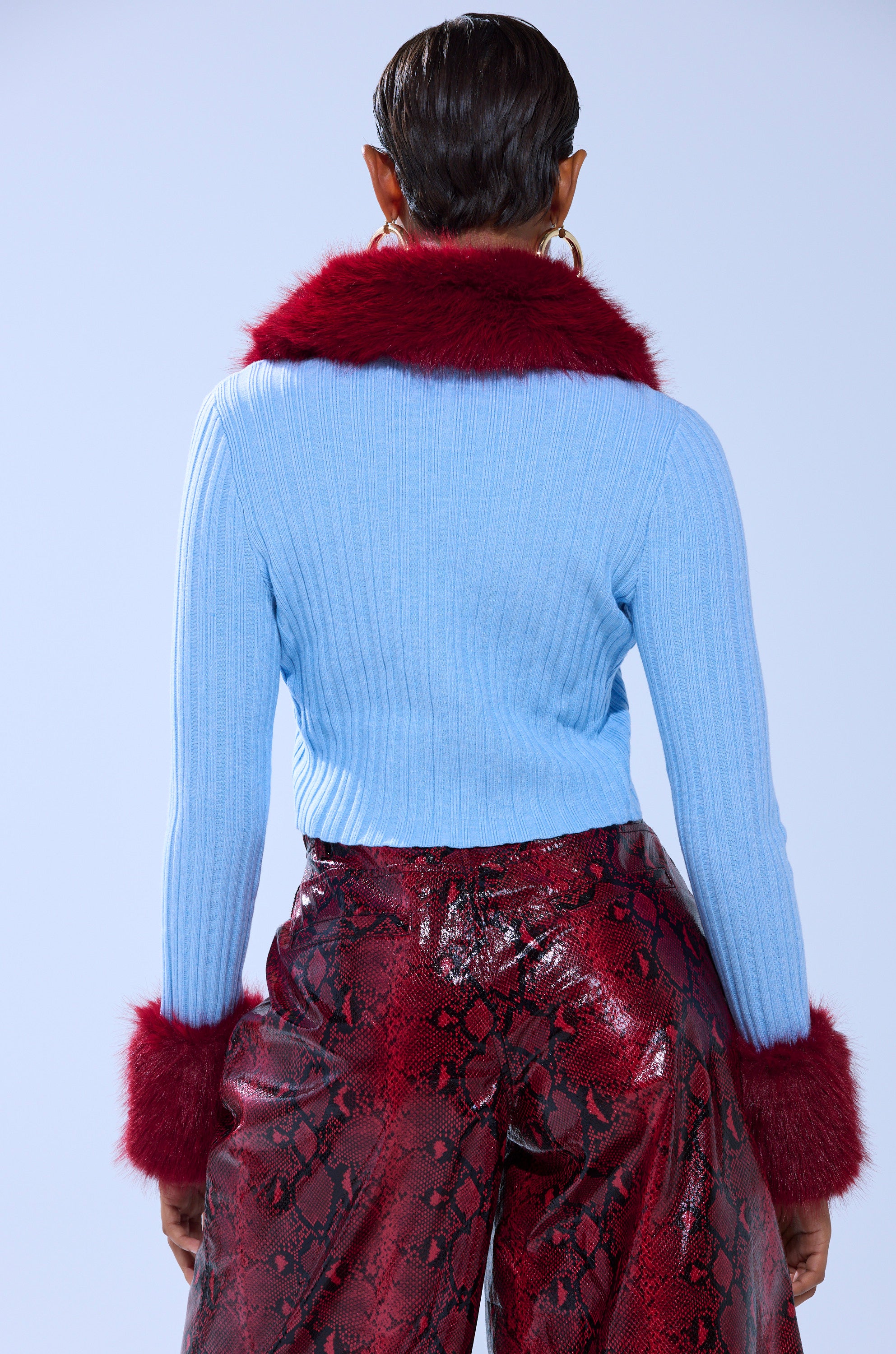 Seen from behind, a person with short dark hair wears the EXTRA IS MY MIDDLE NAME CARDIGAN WITH FUR in light blue, featuring faux fur trim and a cropped fit, paired with shiny red snakeskin-patterned pants.