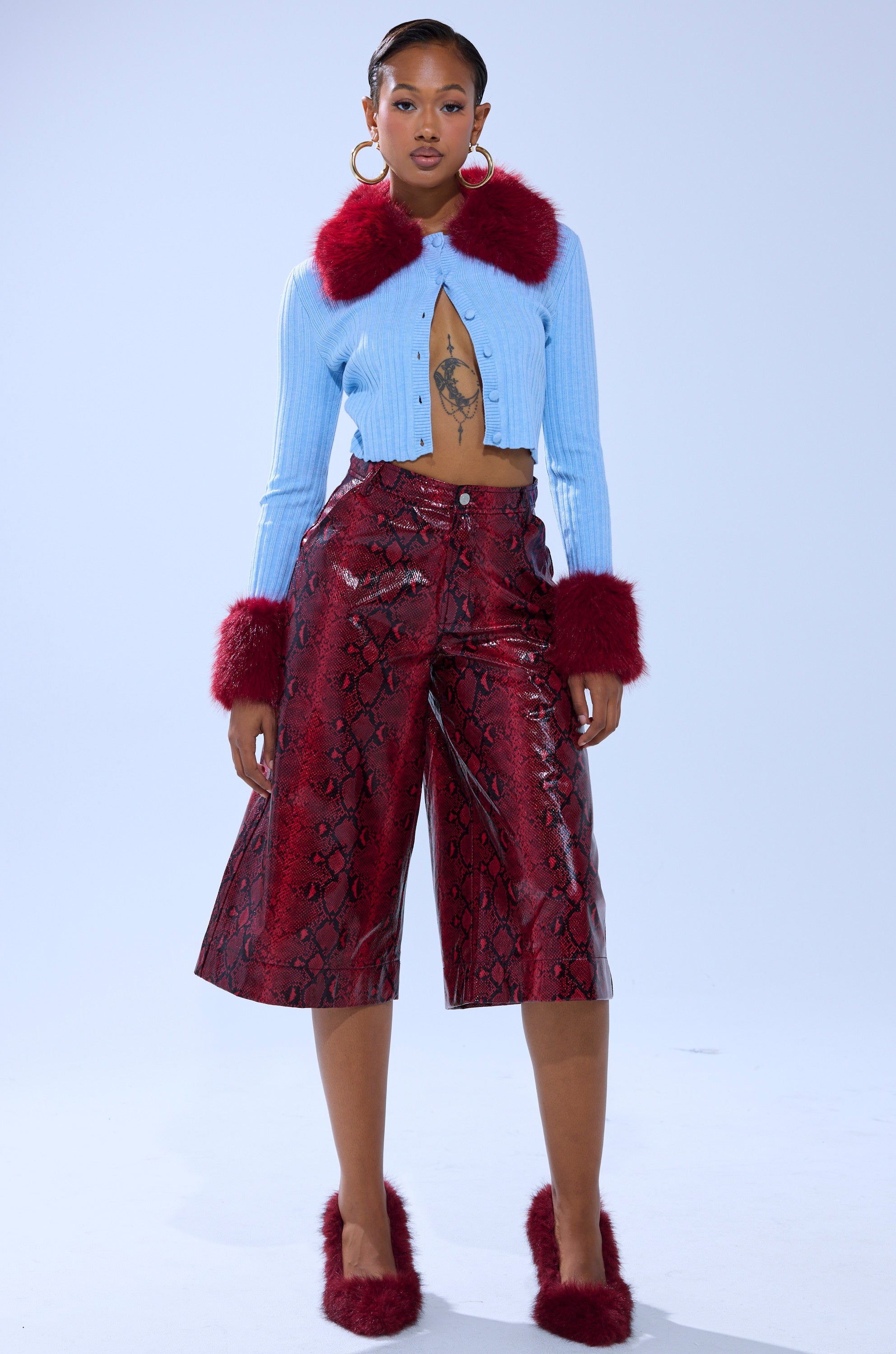 A woman stands confidently wearing the EXTRA IS MY MIDDLE NAME CARDIGAN WITH FUR—a light blue cropped ribbed knit with red faux fur collar and cuffs—paired with red snakeskin culotte pants, red fur slippers, hoop earrings, and a chest tattoo.