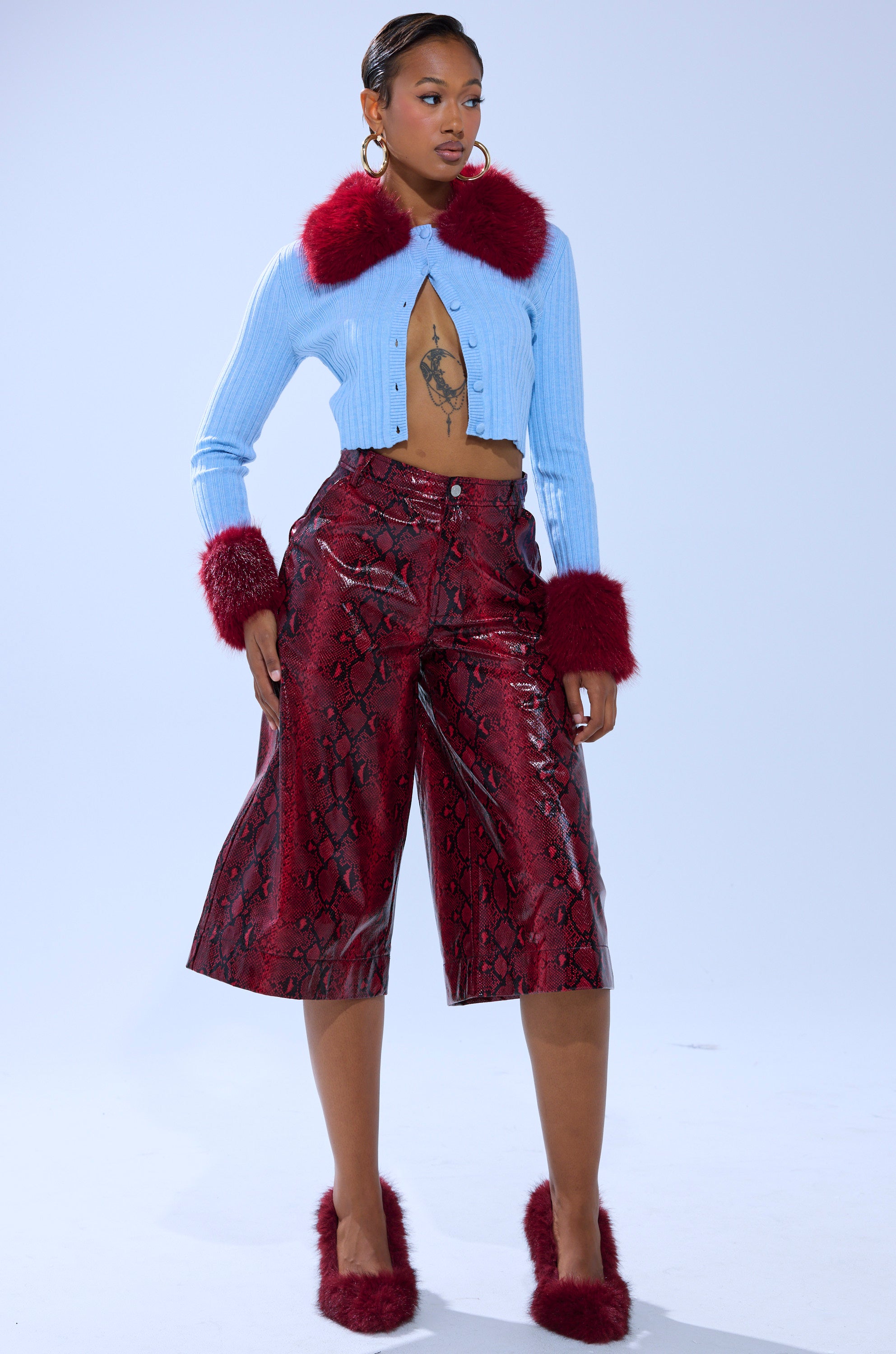 A woman poses wearing the EXTRA IS MY MIDDLE NAME CARDIGAN WITH FUR, featuring a cropped knit design and red faux fur trim, styled with maroon snakeskin culottes, large hoop earrings, and red furry slip-on shoes.