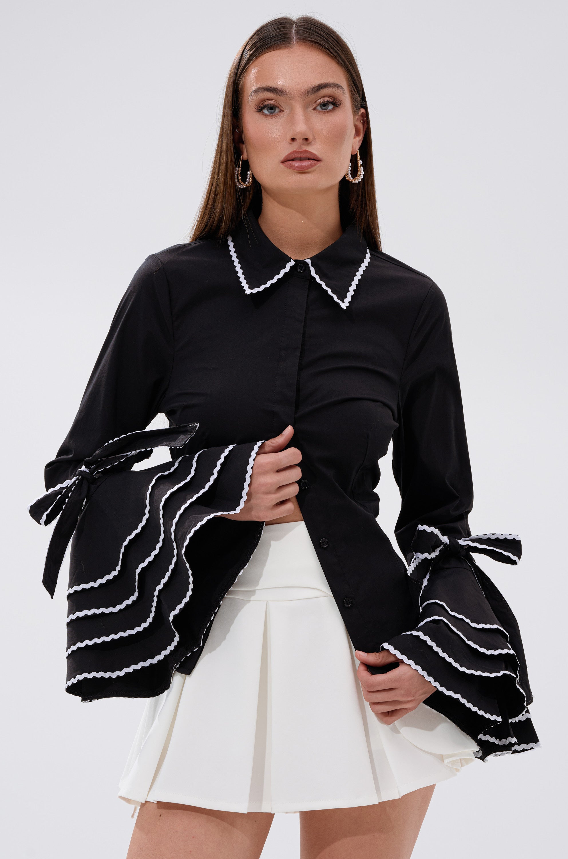 A woman stands against a plain light background wearing the BUSINESS ONLY BUTTON DOWN, a tailored black business shirt with white-trimmed flared bell sleeves, paired with a white pleated skirt.