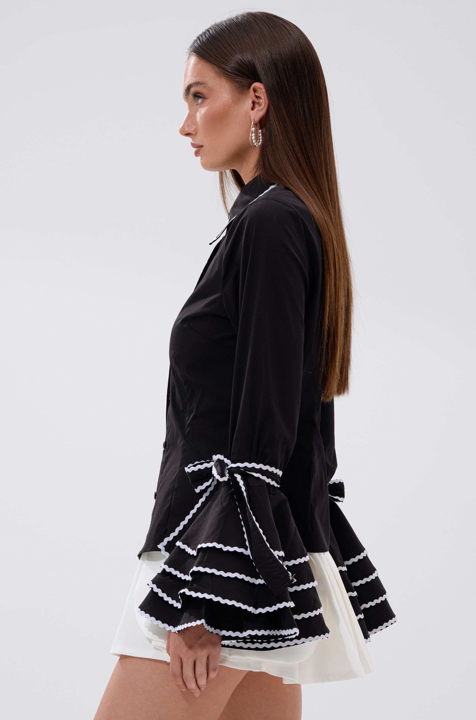 A woman stands in profile against a plain background wearing the BUSINESS ONLY BUTTON DOWN, a black tailored shirt with white-trimmed, flared ruffled bell sleeves, paired with a white skirt. She has long brown hair and hoop earrings.