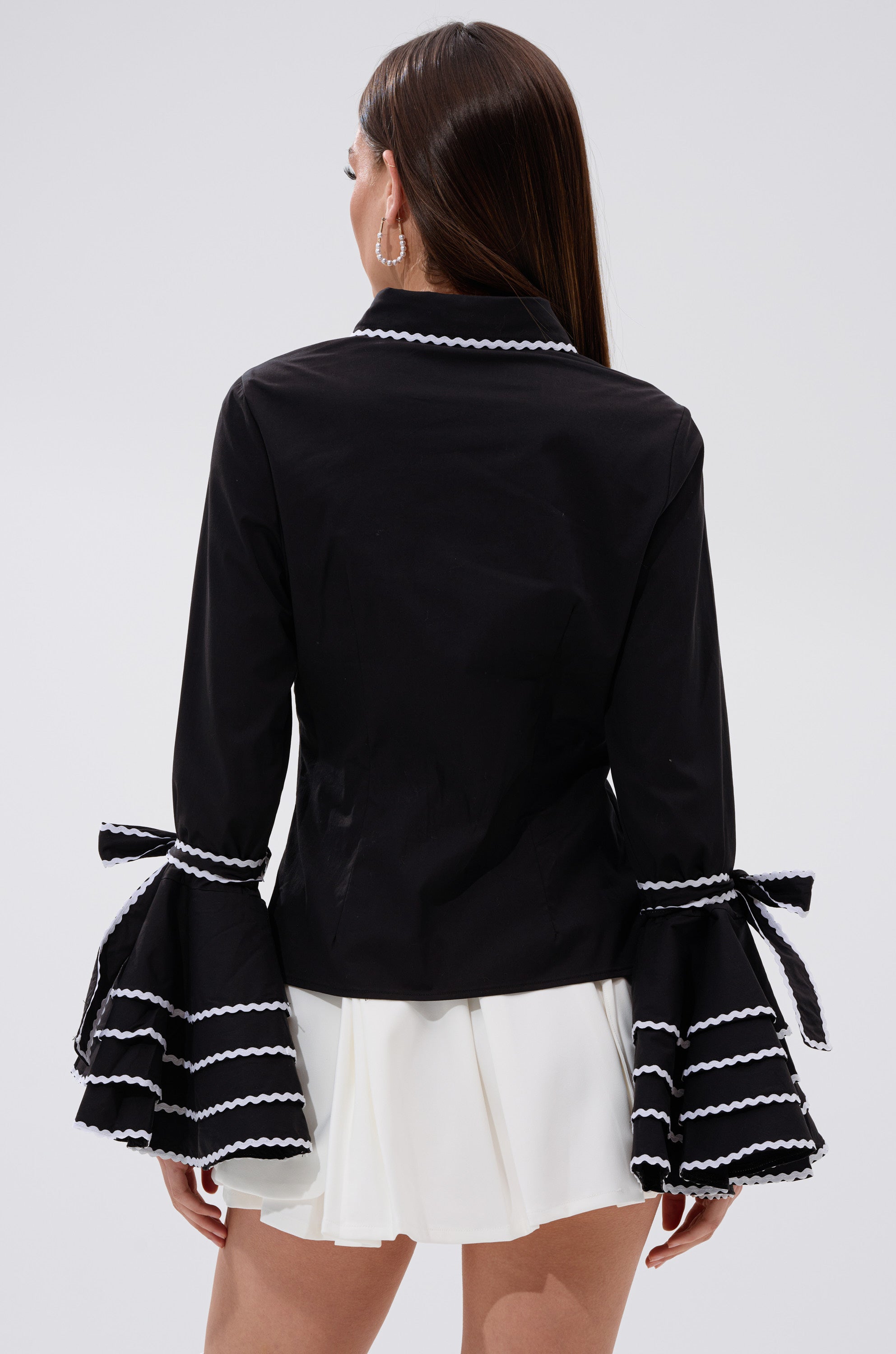 A woman is shown from behind wearing the BUSINESS ONLY BUTTON DOWN, a black jacket with flared bell sleeves featuring white stripe trim and decorative bows, paired with a short white skirt.