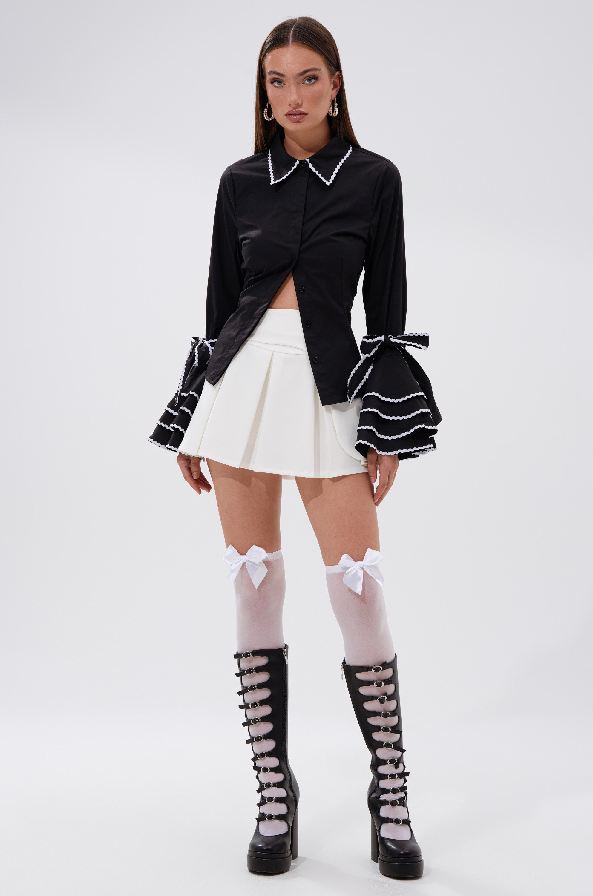 A woman stands against a plain background wearing the BUSINESS ONLY BUTTON DOWN, a black tailored shirt with ruffled sleeves, styled with a white pleated skirt, white knee-high socks with bows, and black platform lace-up boots.