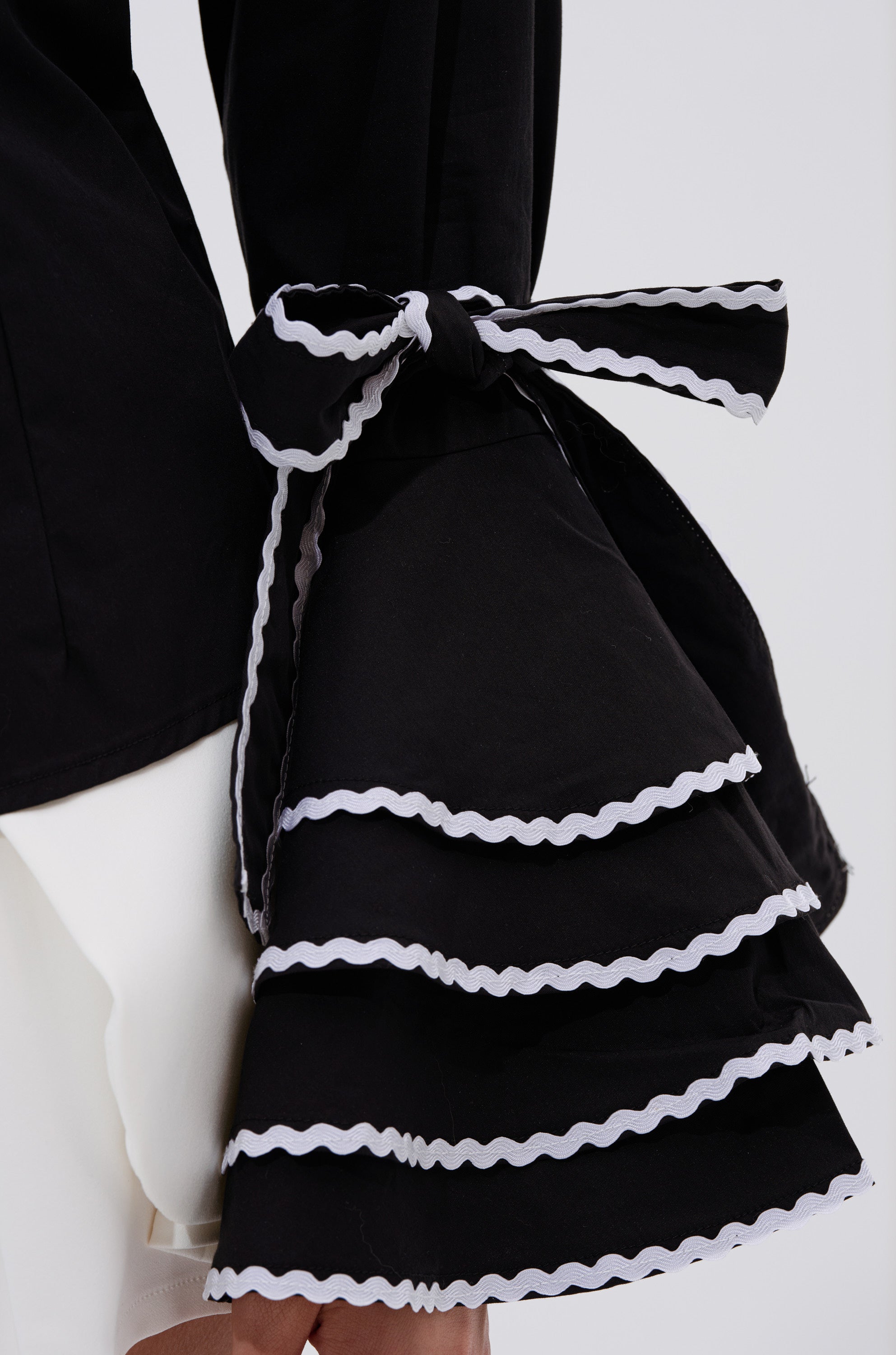 A close-up of a person wearing the BUSINESS ONLY BUTTON DOWN, a black tailored shirt featuring tiered, flared bell sleeves trimmed in white and tied with a matching ribbon, contrasting elegantly with crisp white fabric underneath.