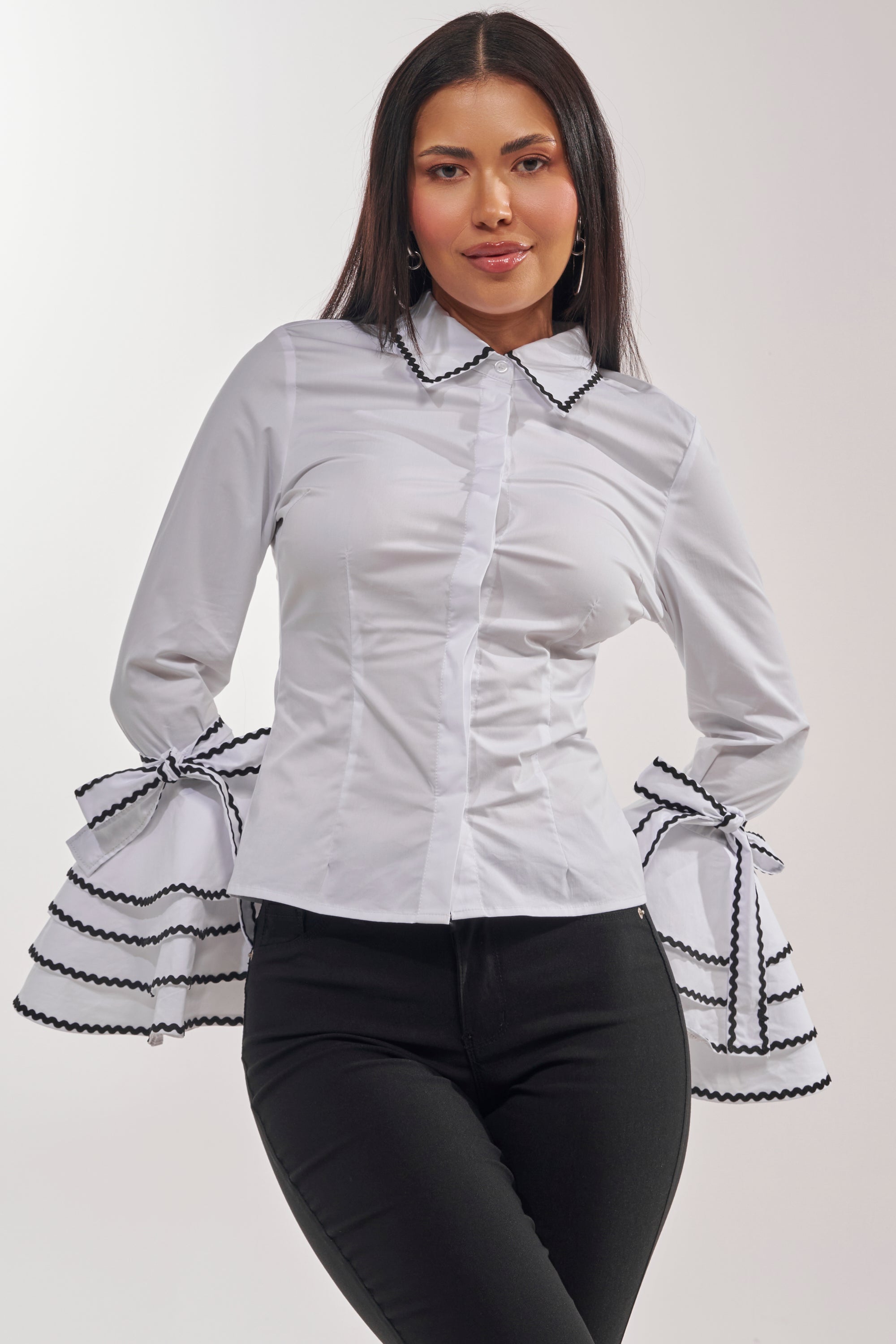 A woman stands against a plain white background, smiling slightly with her hands on her hips, wearing the BUSINESS ONLY BUTTON DOWN IN WHITE featuring large ruffled bell sleeves with contrast trim and paired with black pants. 3