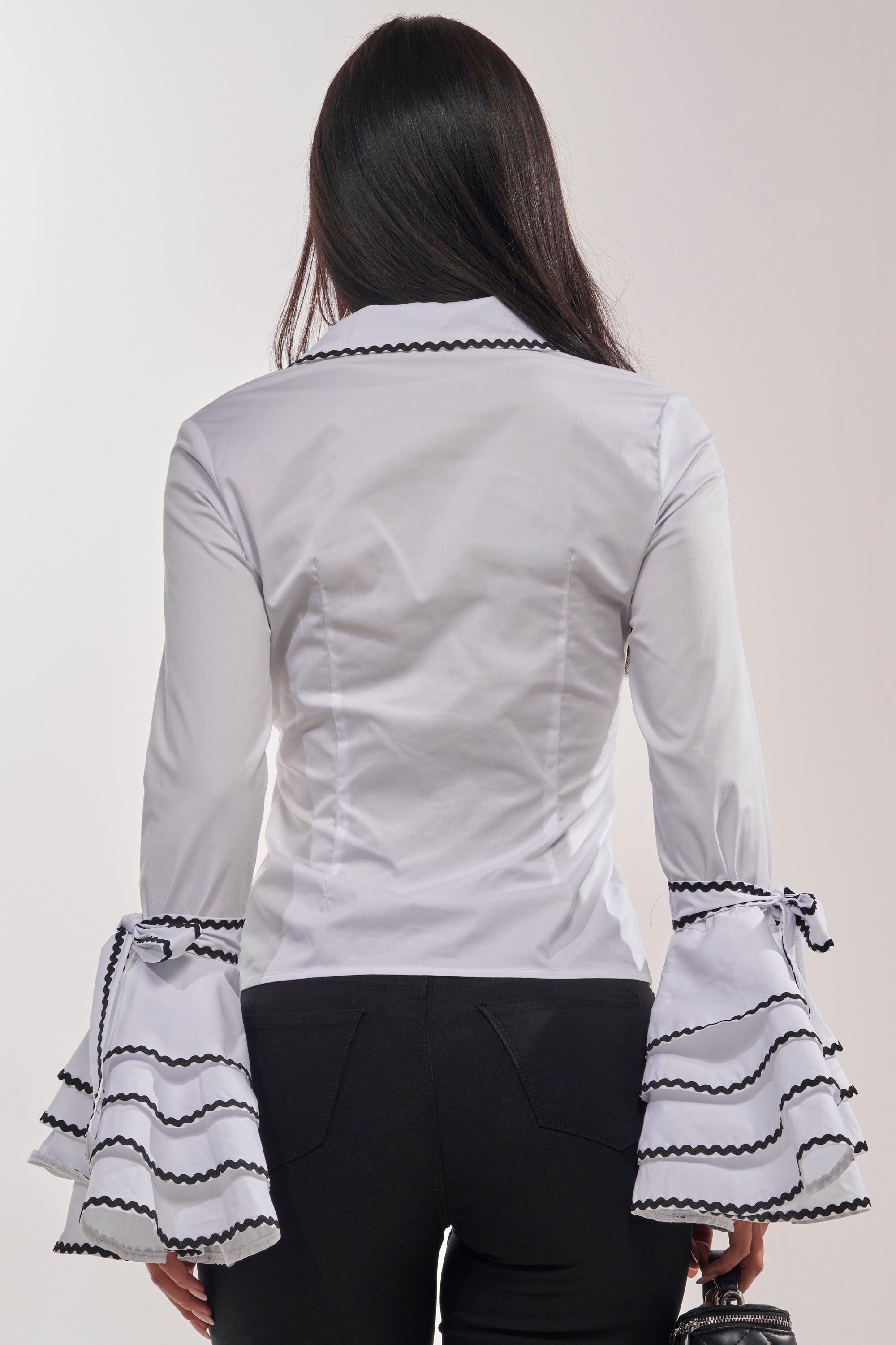 A woman with long dark hair faces away, wearing the BUSINESS ONLY BUTTON DOWN IN WHITE—a fitted blouse with black contrast trim—paired with black pants and holding a small purse in her left hand. 5