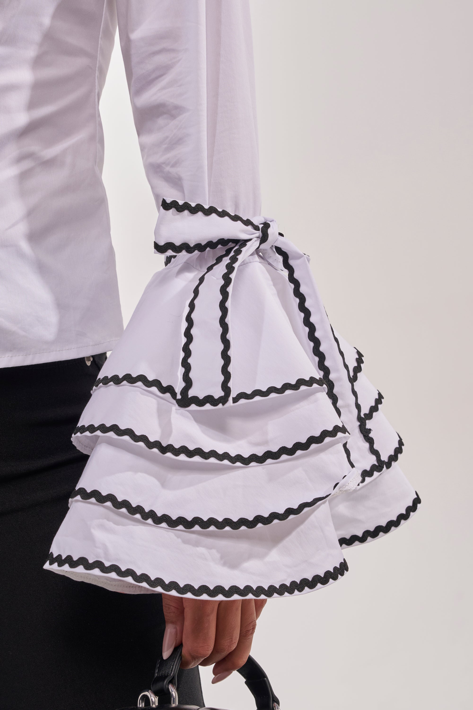 A close-up of a person wearing the BUSINESS ONLY BUTTON DOWN IN WHITE, featuring dramatic, layered ruffled sleeves with black trim, paired with a black skirt and holding a small black handbag. 8