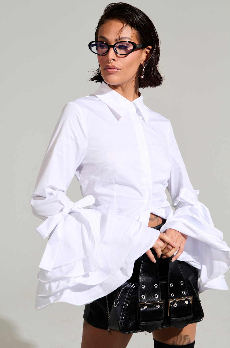 A confident individual with short, wet hair and glasses poses wearing the VANJA BUTTON DOWN BLOUSE, characterized by its oversized bell sleeves. They pair it with a black skirt featuring silver buckles, standing against a neutral gray backdrop.