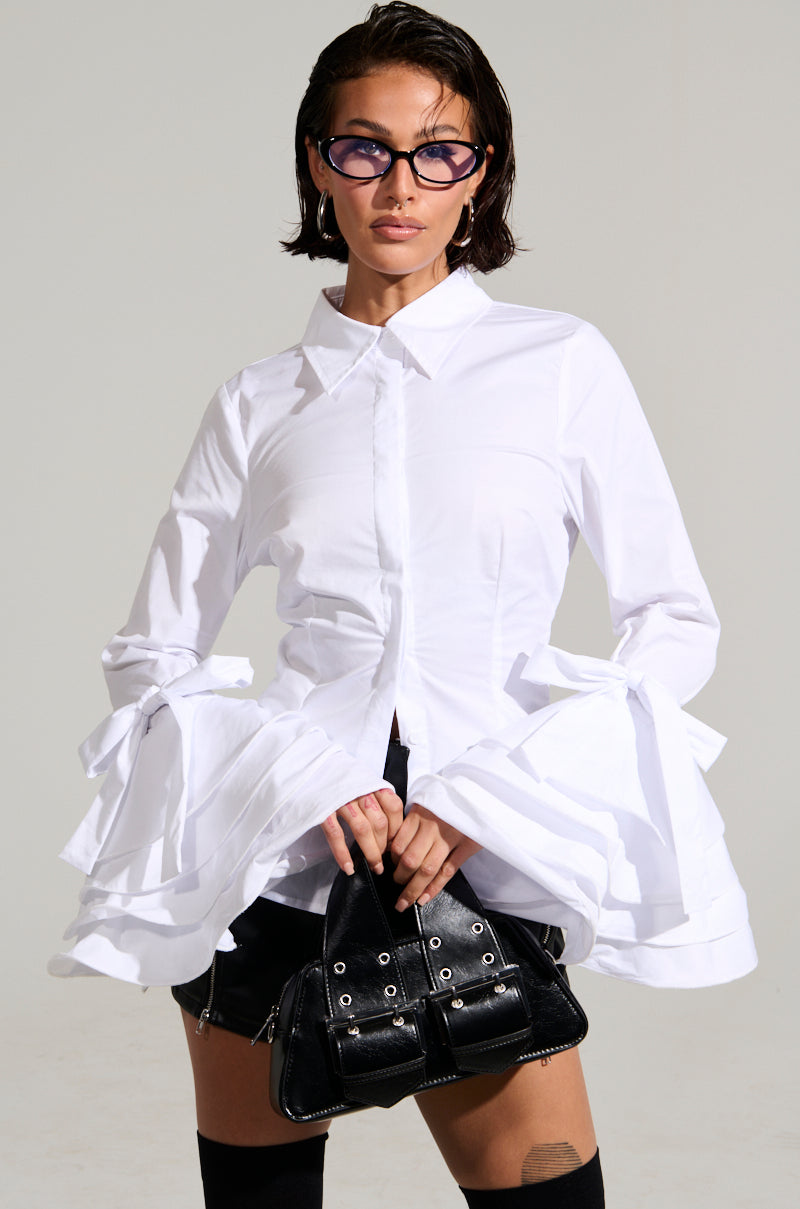 Dressed confidently in the VANJA BUTTON DOWN BLOUSE, which features oversized bell sleeves in crisp poplin fabric, they pair it with black shorts. Holding a black handbag and wearing dark rectangular glasses, they stand against a plain light gray background.