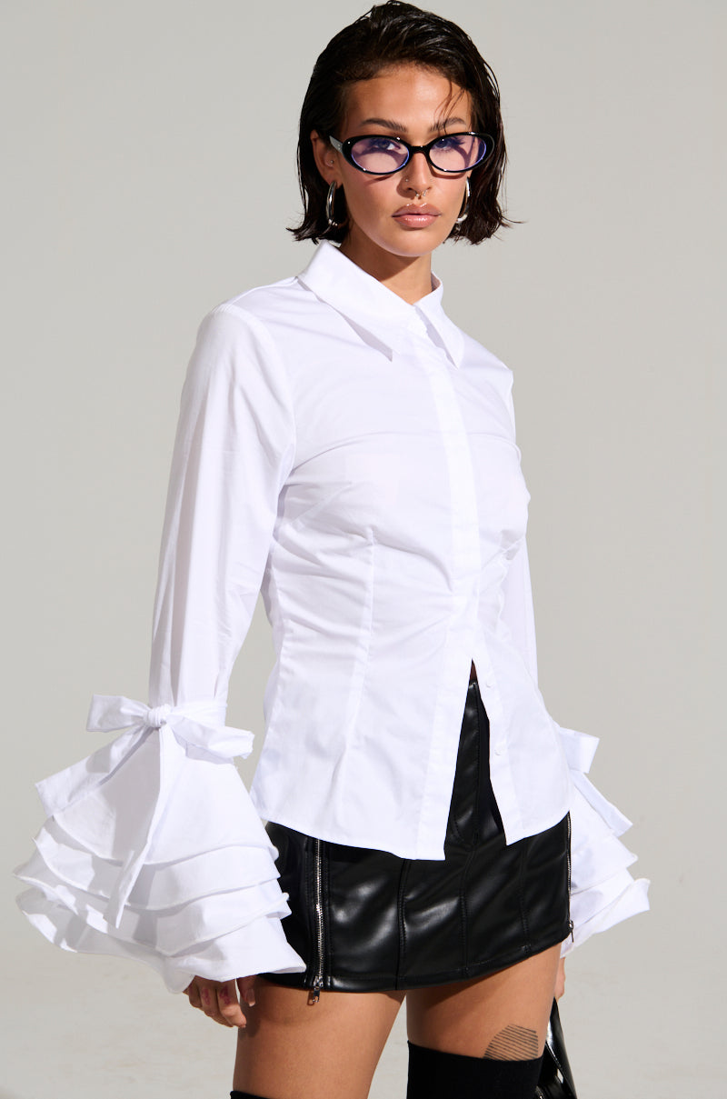A person wearing the VANJA BUTTON DOWN BLOUSE, featuring elegant tiered ruffle sleeves, paired with a sleek black leather skirt. The poplin fabric enhances their modern look. With short dark hair and black-rimmed glasses, they strike a confident pose against a plain background.