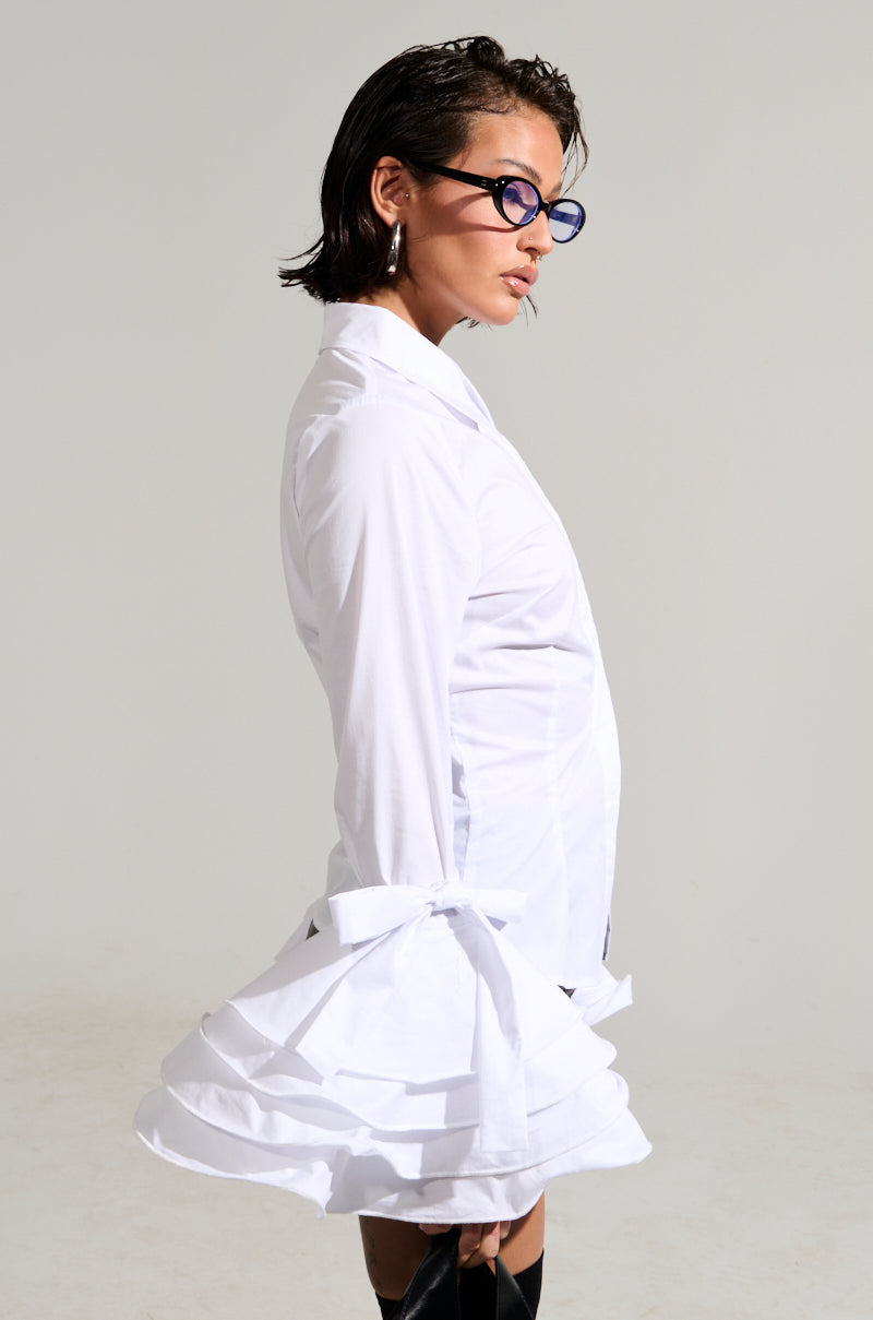A person with short dark hair and blue sunglasses poses against a plain background, gazing to the side while wearing a white VANJA BUTTON DOWN BLOUSE that features ruffled, ribbon-tied sleeves made from poplin fabric.