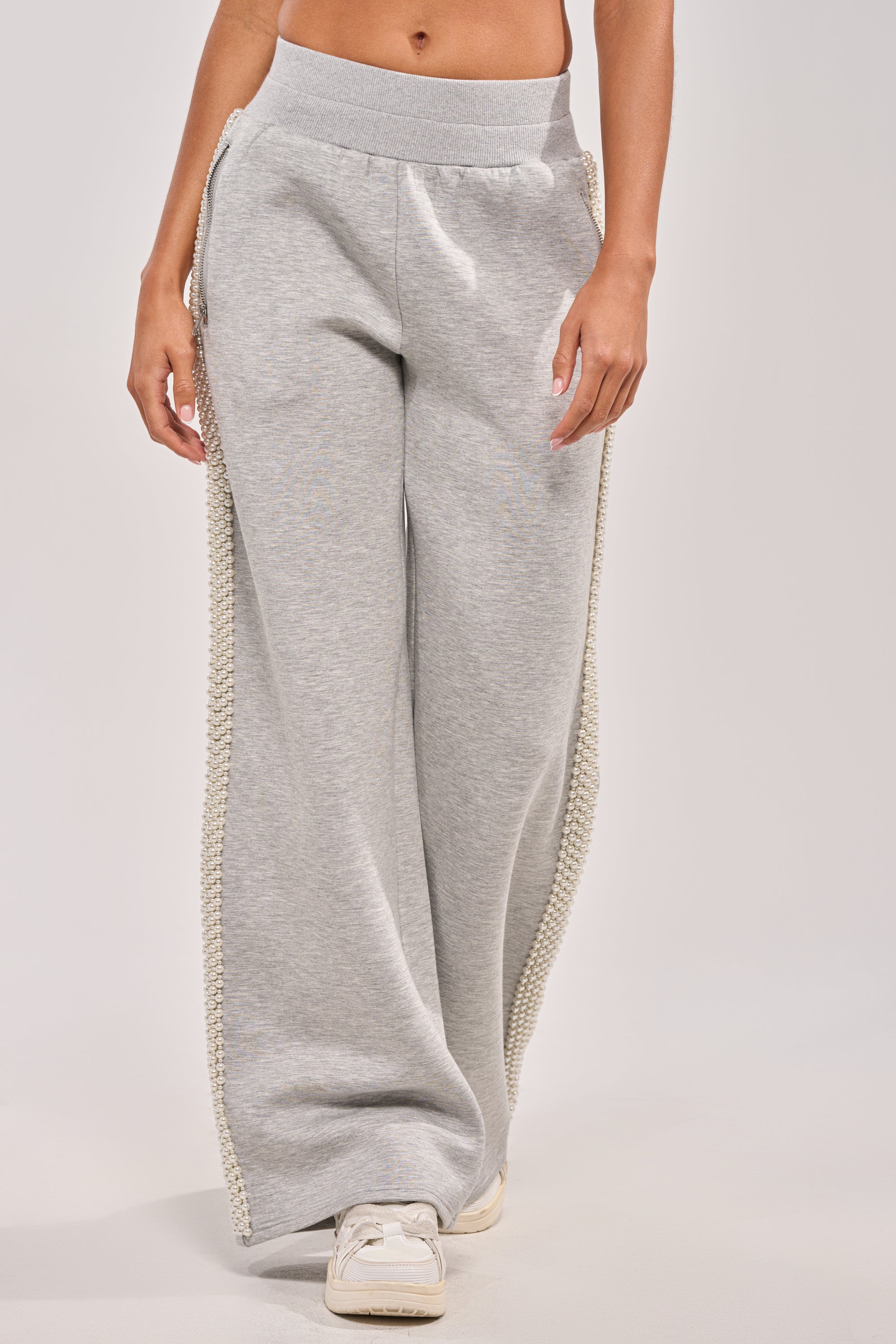 Someone wears PEARL GIRL WIDE LEG JOGGER IN GREY featuring a faux pearl trim on the side seams and an elasticated waistband, styled with white sneakers. The person's upper body is not shown. 1