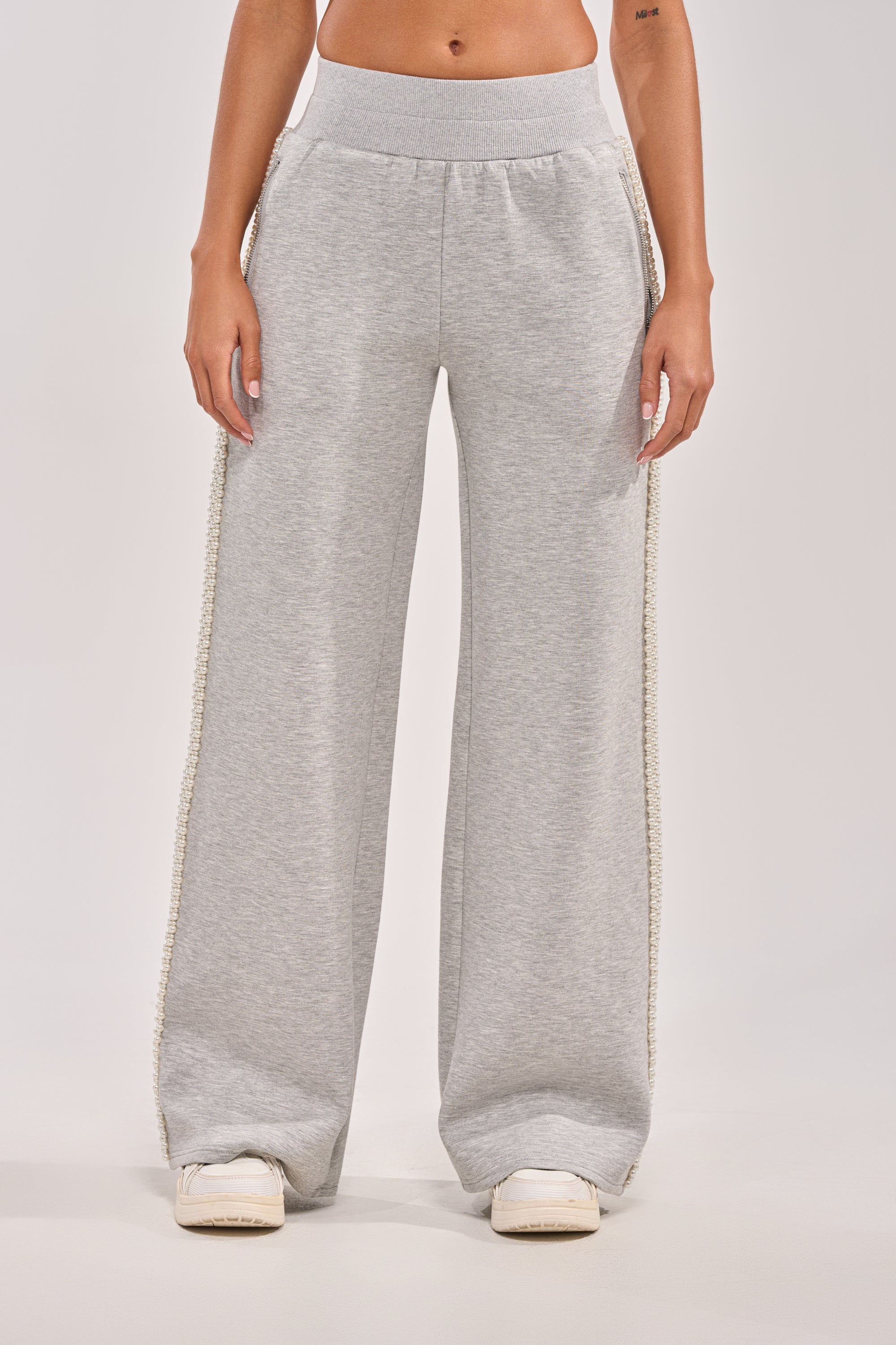 A person showcases the PEARL GIRL WIDE LEG JOGGER IN GREY, featuring faux pearl trim on the sides, paired with white sneakers against a plain background. Only the lower half of their body is visible. 2