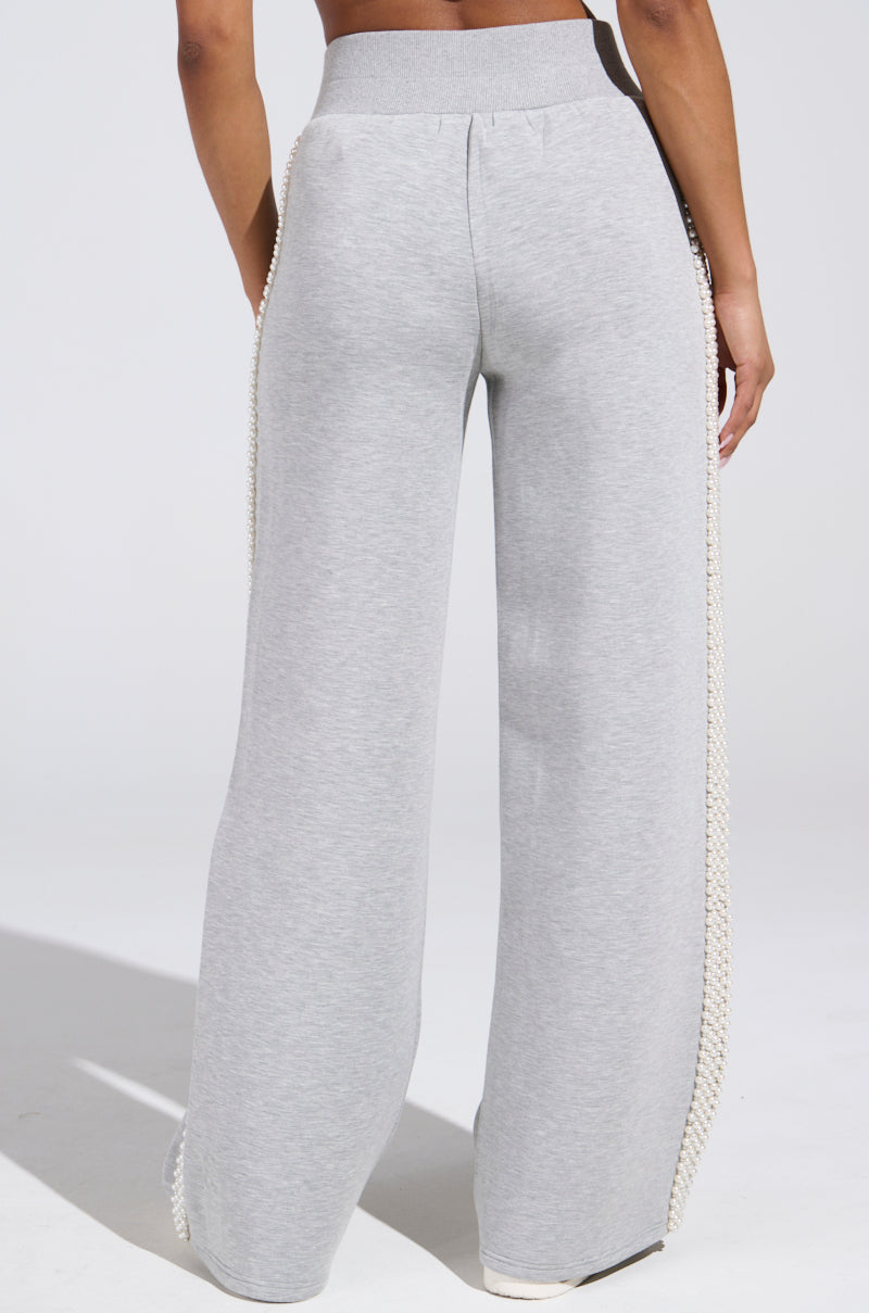 A person wearing PEARL GIRL WIDE LEG JOGGER IN GREY, with elegant white crochet along the side seams, stands against a white background. The athleisure style image highlights the back view of these chic pants.