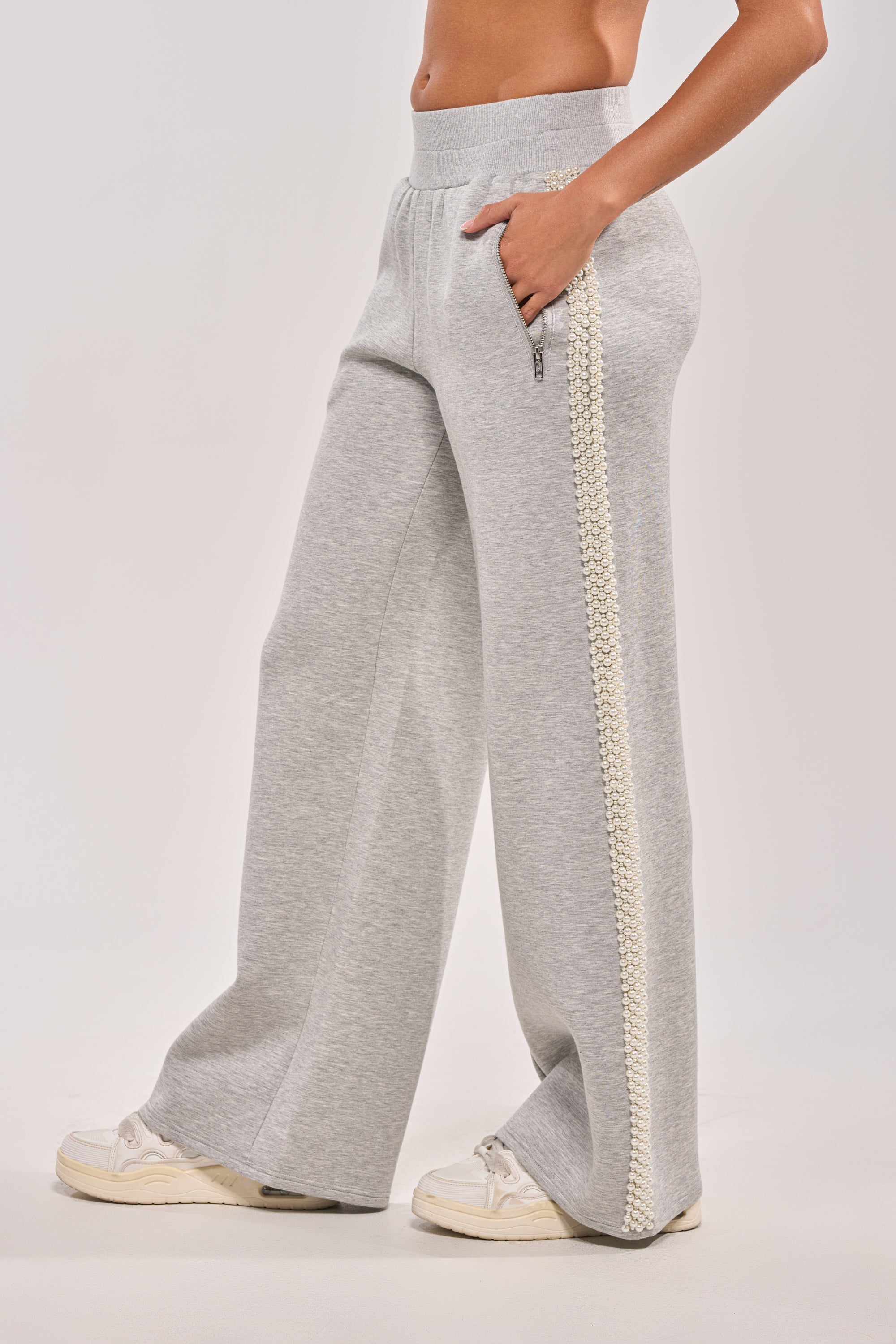 A person models the PEARL GIRL WIDE LEG JOGGER IN GREY, featuring a white braided side stripe, with one hand in the pocket and paired with white sneakers against a plain light background. 4
