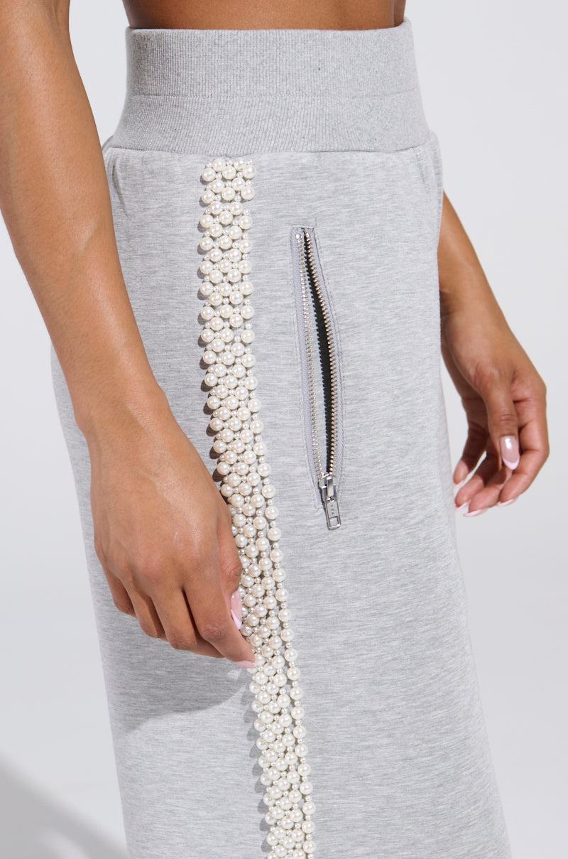 A person sports the PEARL GIRL WIDE LEG JOGGER IN GREY, featuring pearl details along the side seam for an athleisure chic vibe. A zippered pocket enhances functionality, while light pink nail polish accents their look, complemented by a partially visible crop top.