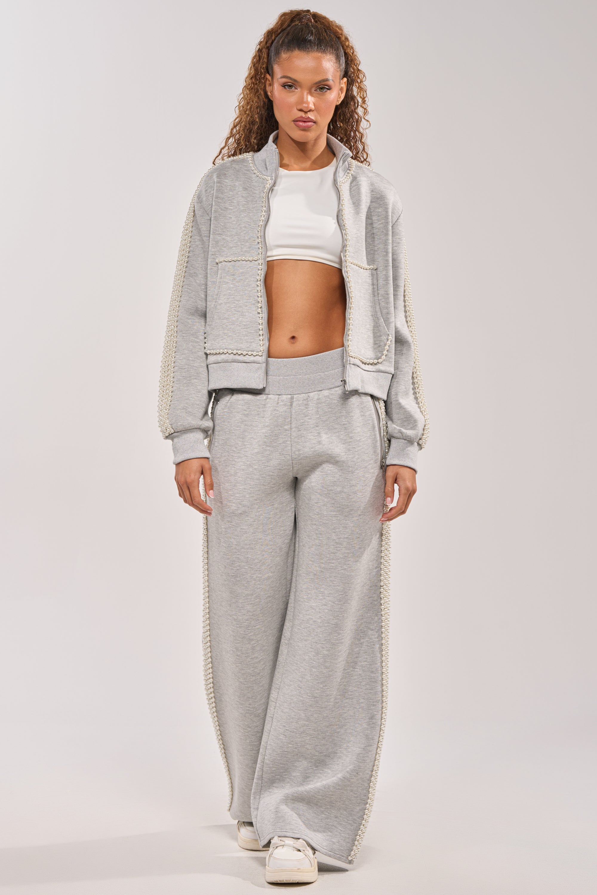 A woman stands against a neutral background wearing the PEARL GIRL WIDE LEG JOGGER IN GREY, paired with a light gray zip-up jacket, white crop top, and white sneakers, showcasing a relaxed silhouette. 7