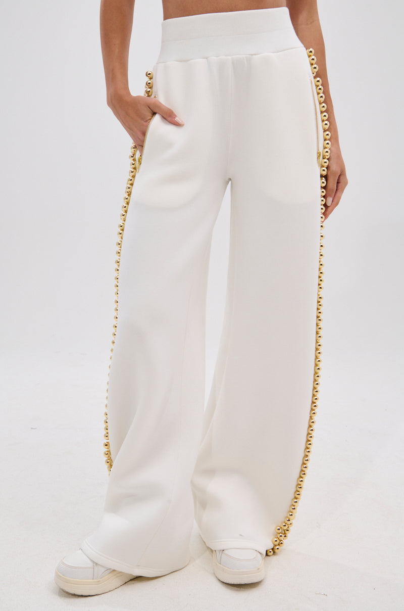 A person wears the BALLS TO THE WALL JOGGER IN WHITE with metallic gold-tone ball embellishments and a high-rise elastic waistband, paired with white sneakers and a cropped white top, standing against a plain white background.