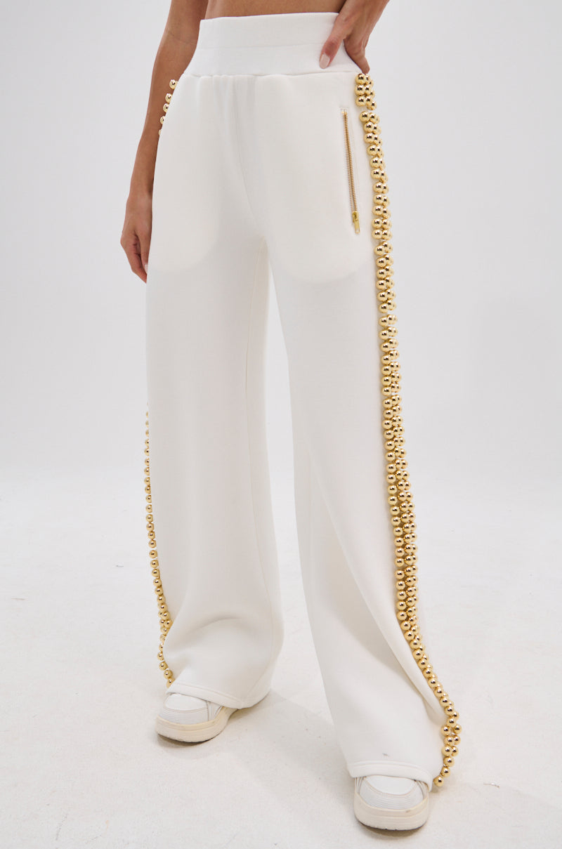 A person models the BALLS TO THE WALL JOGGER IN WHITE—high-waisted, wide-leg pants with metallic gold-tone ball details along the side seams—worn with white sneakers. Only their lower half is shown against a plain background.