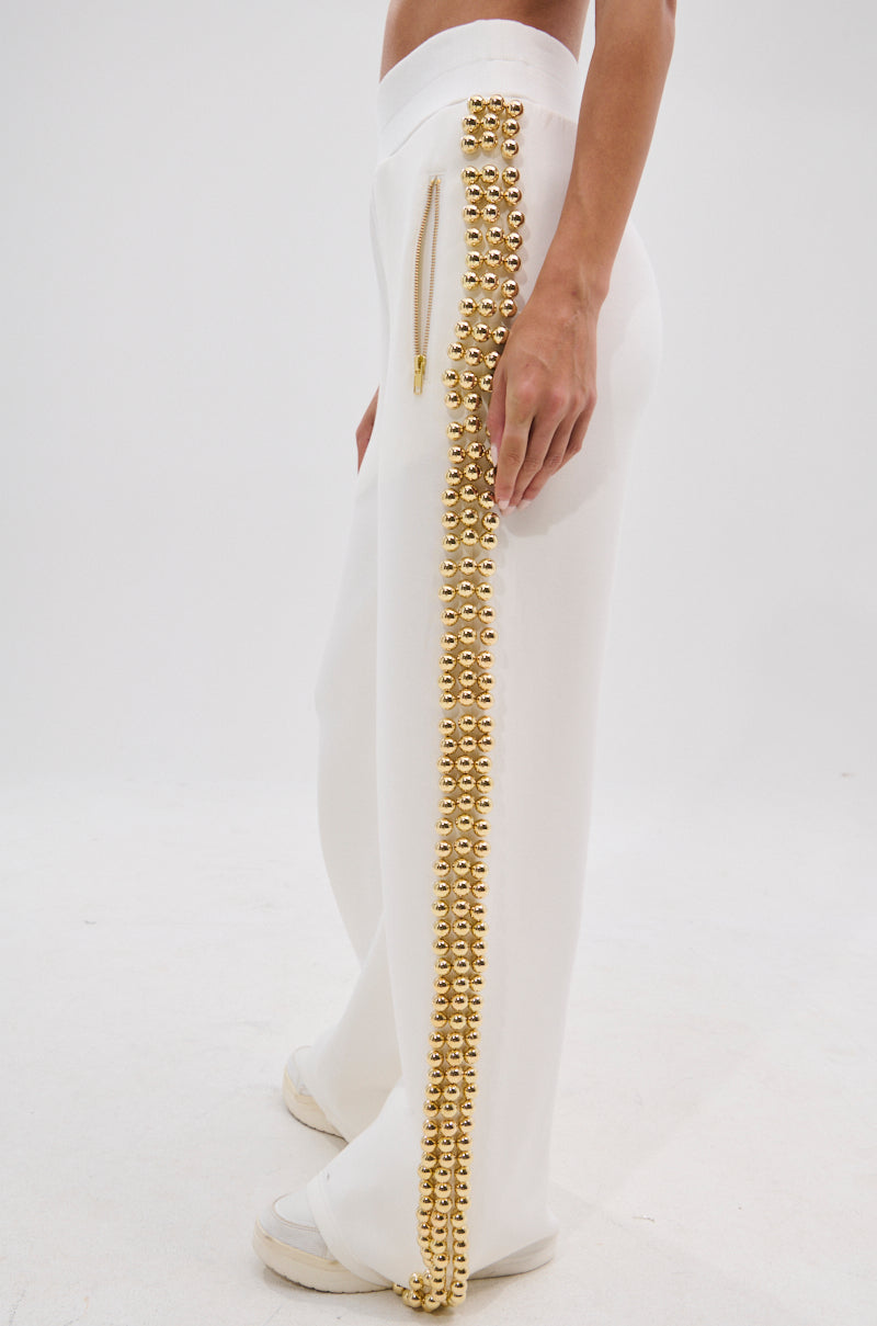 Wearing the BALLS TO THE WALL JOGGER IN WHITE—white joggers featuring metallic gold-tone ball details on the side seams and a high-rise elastic waistband—paired with white sneakers, the person stands against a plain white background.