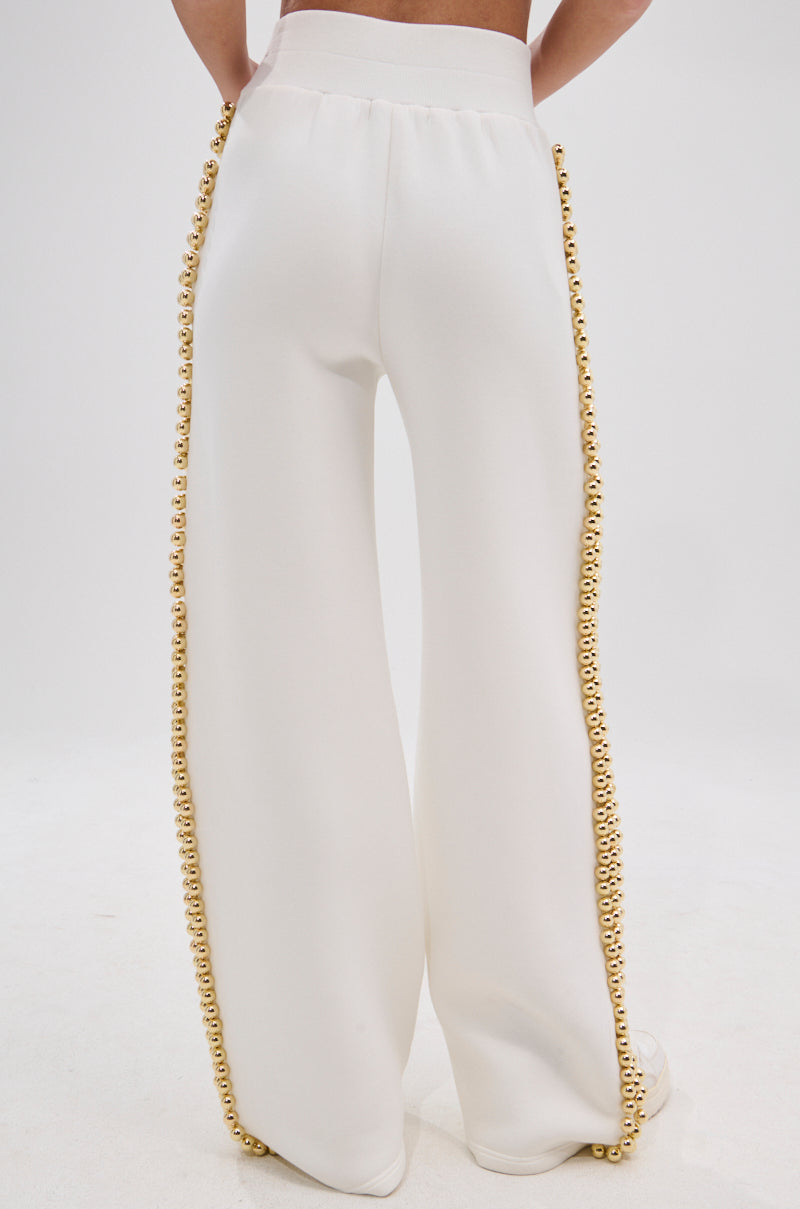 Someone models the BALLS TO THE WALL JOGGER IN WHITE—high-waisted, wide-leg joggers with a high-rise elastic waistband and metallic gold ball accents along the side seams—against a plain white background.